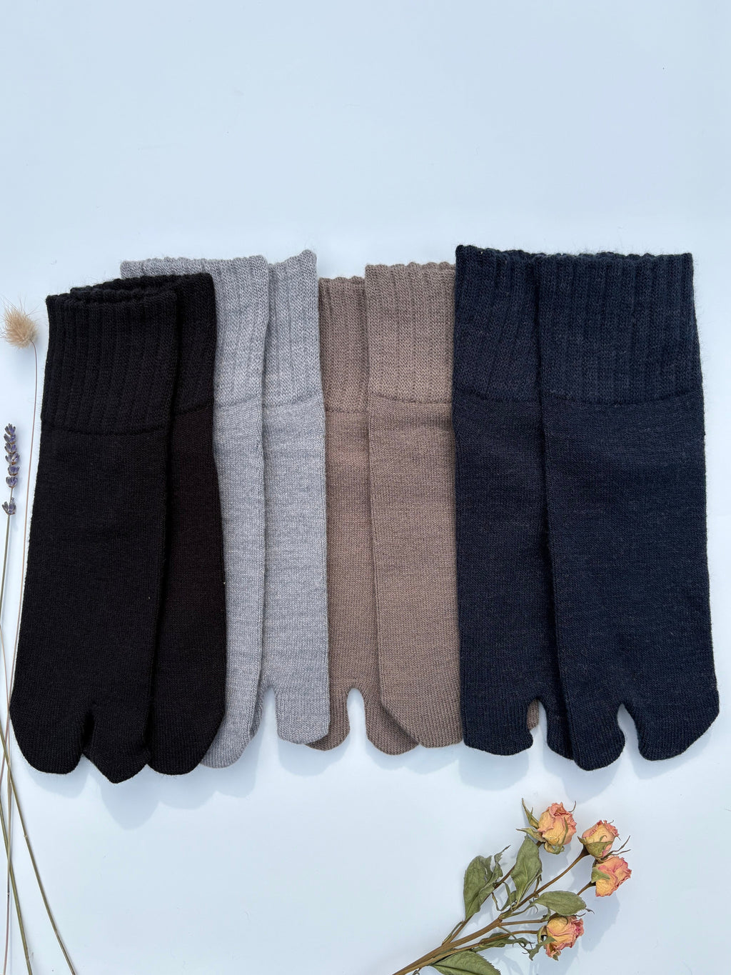 Four pairs of plain tabi split-toe socks lined up in grey, dark brown, taupe, and navy — soft ribbed-cuff Japanese tabi socks in neutral tones styled with dried flowers. Wool blend super soft