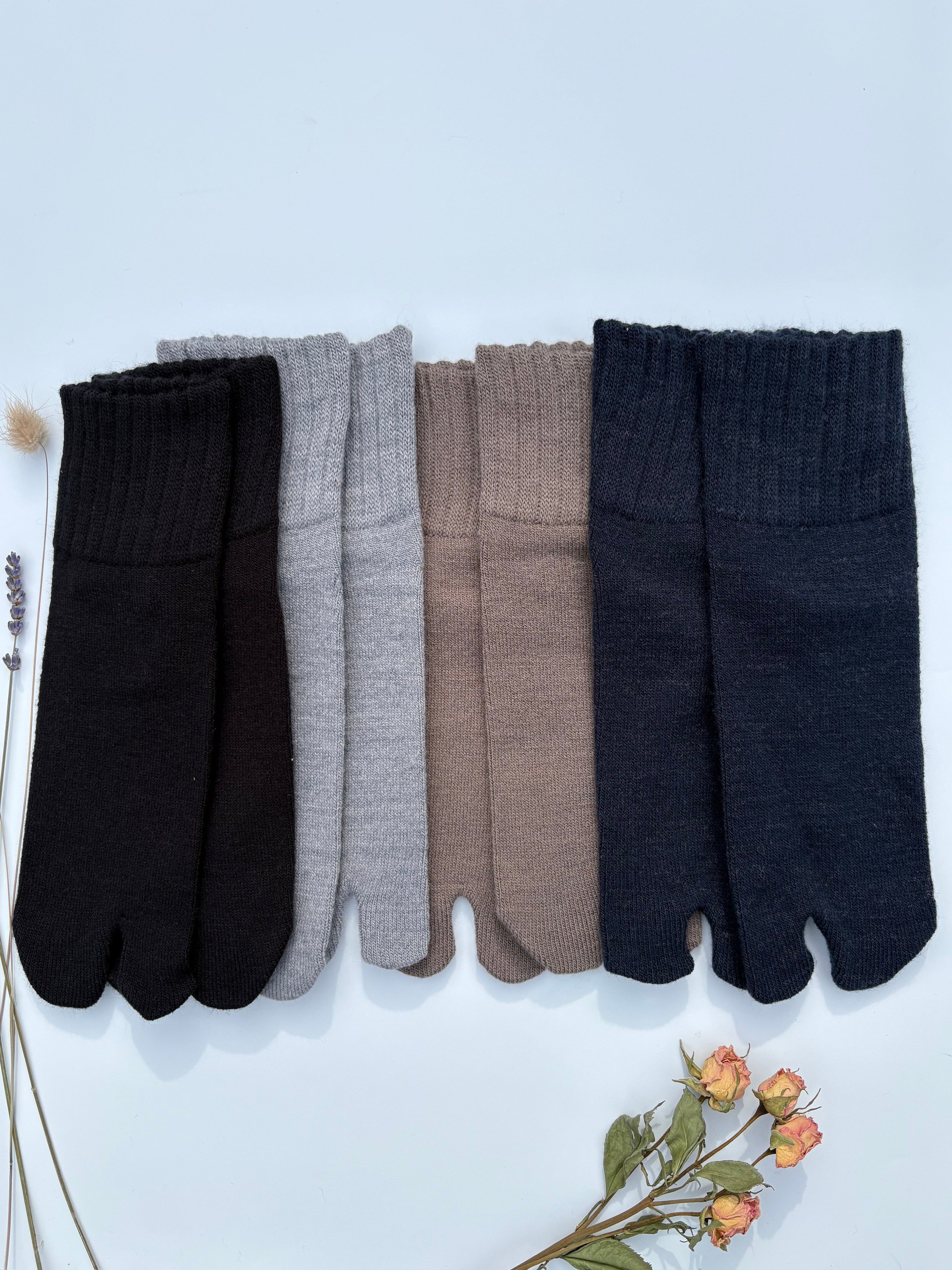 Four pairs of plain tabi split-toe socks lined up in grey, dark brown, taupe, and navy — soft ribbed-cuff Japanese tabi socks in neutral tones styled with dried flowers. Wool blend super soft