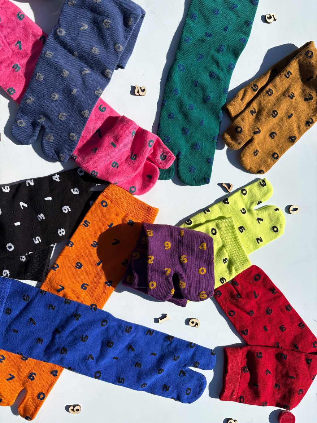Flat lay of tabi split-toe socks in number print across eight colourways including orange, blue, purple, pink, black, red, yellow-green, and tan — styled with wooden number props