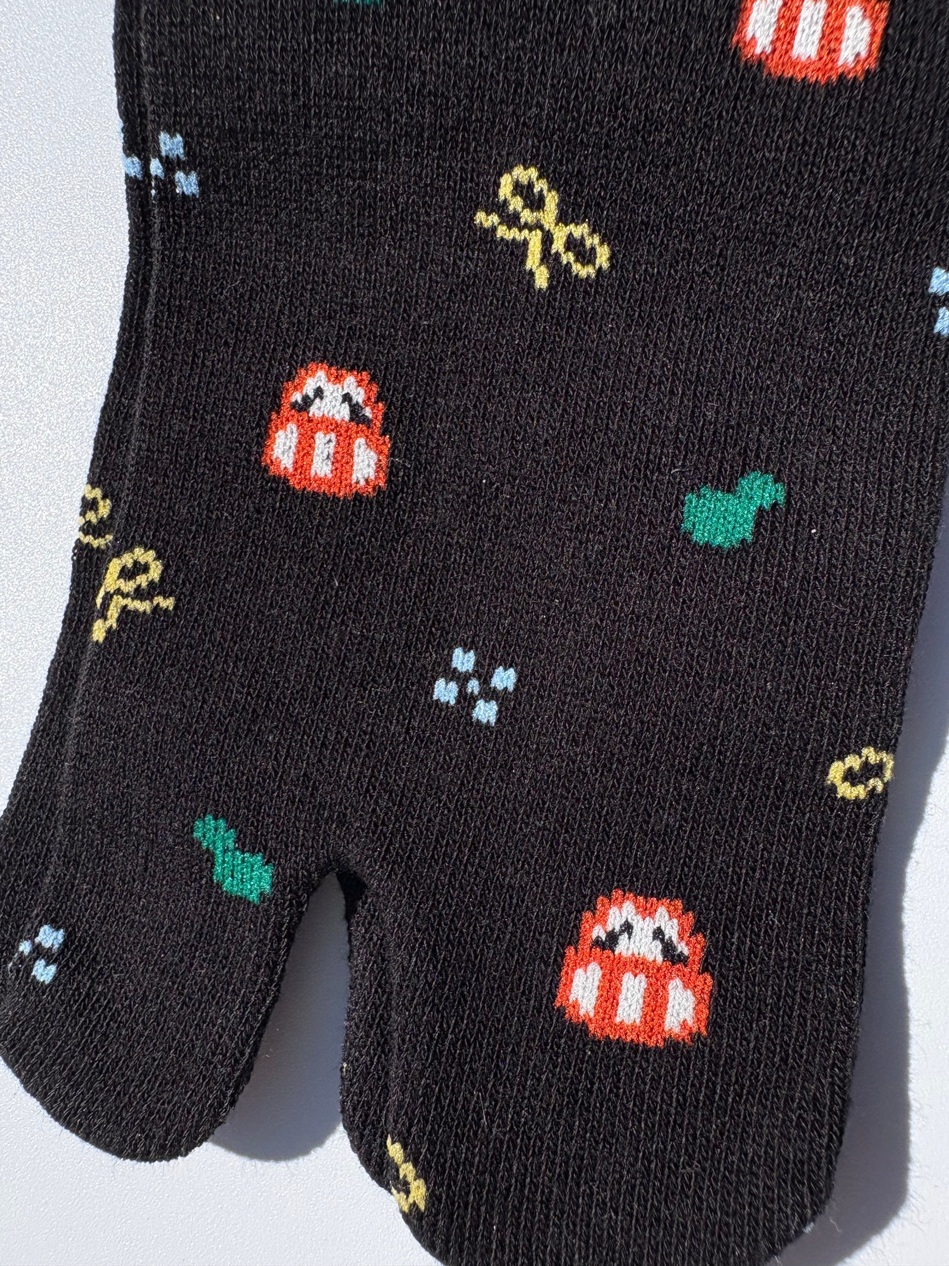 Close-up of black tabi split-toe sock toe showing pixel art daruma doll, green cactus, and paw print motifs — Japanese novelty lucky charm tabi socks