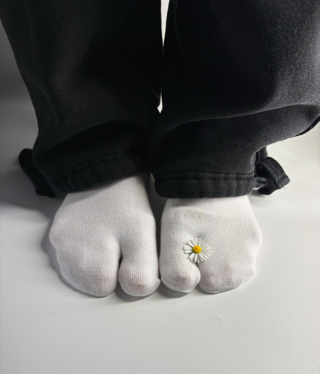 White tabi split-toe socks worn on feet with a single real daisy flower resting between the toes — styled with black wide-leg trousers — minimalist Japanese tabi socks