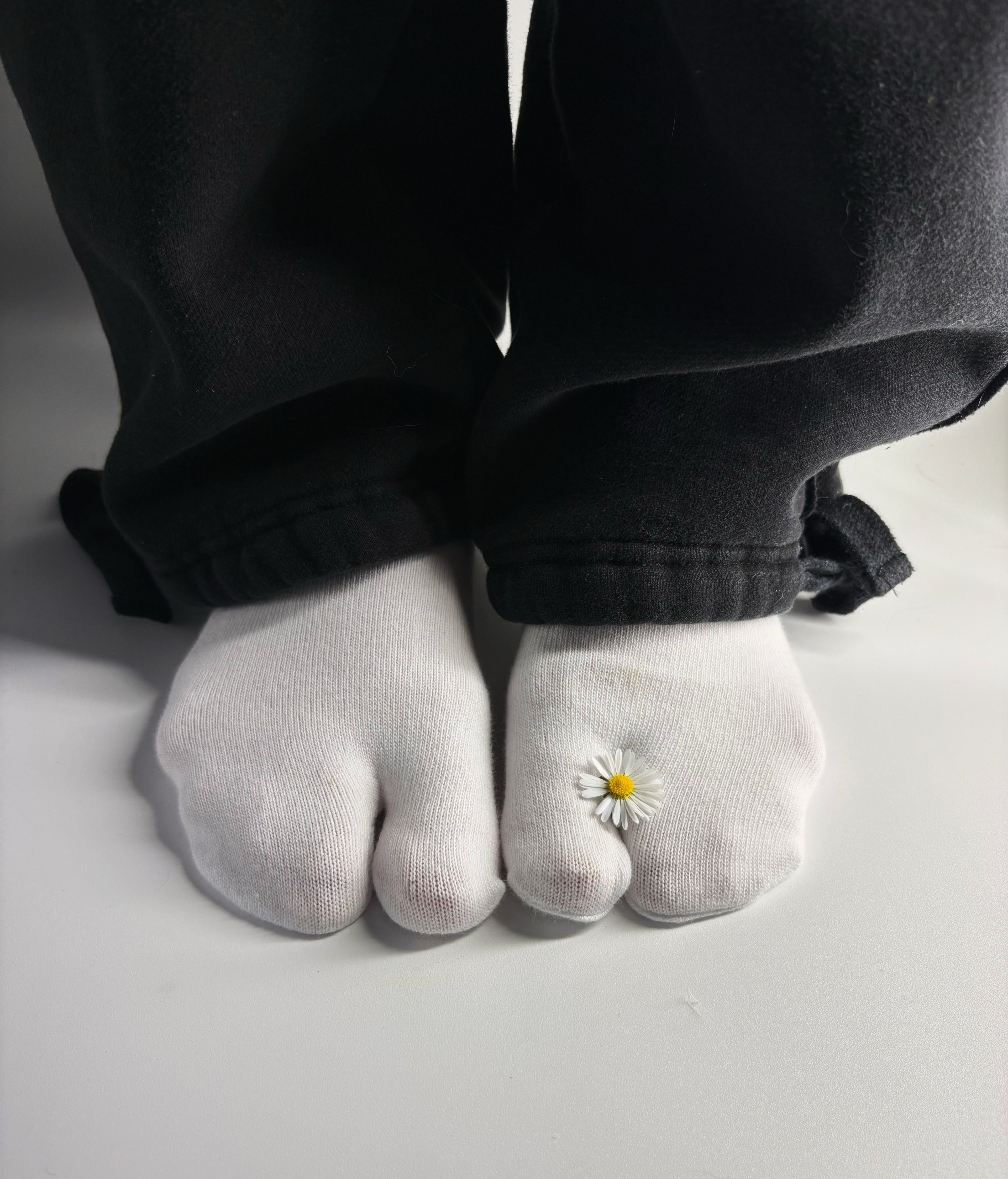 White tabi split-toe socks worn on feet with a single real daisy flower resting between the toes — styled with black wide-leg trousers — minimalist Japanese tabi socks
