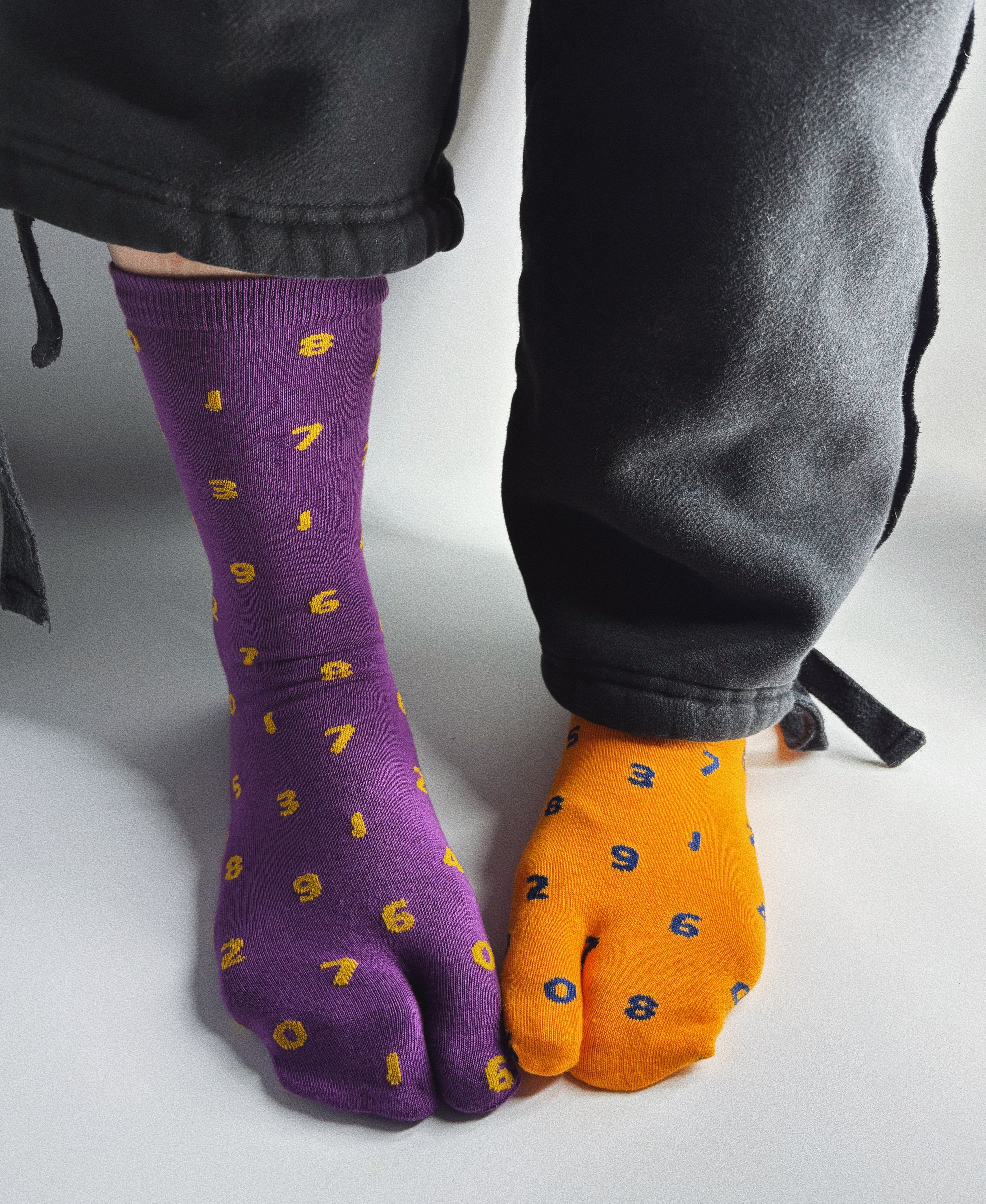 Mismatched tabi split-toe socks worn on feet — purple with yellow number print and orange with navy number print — styled with grey wide-leg trousers