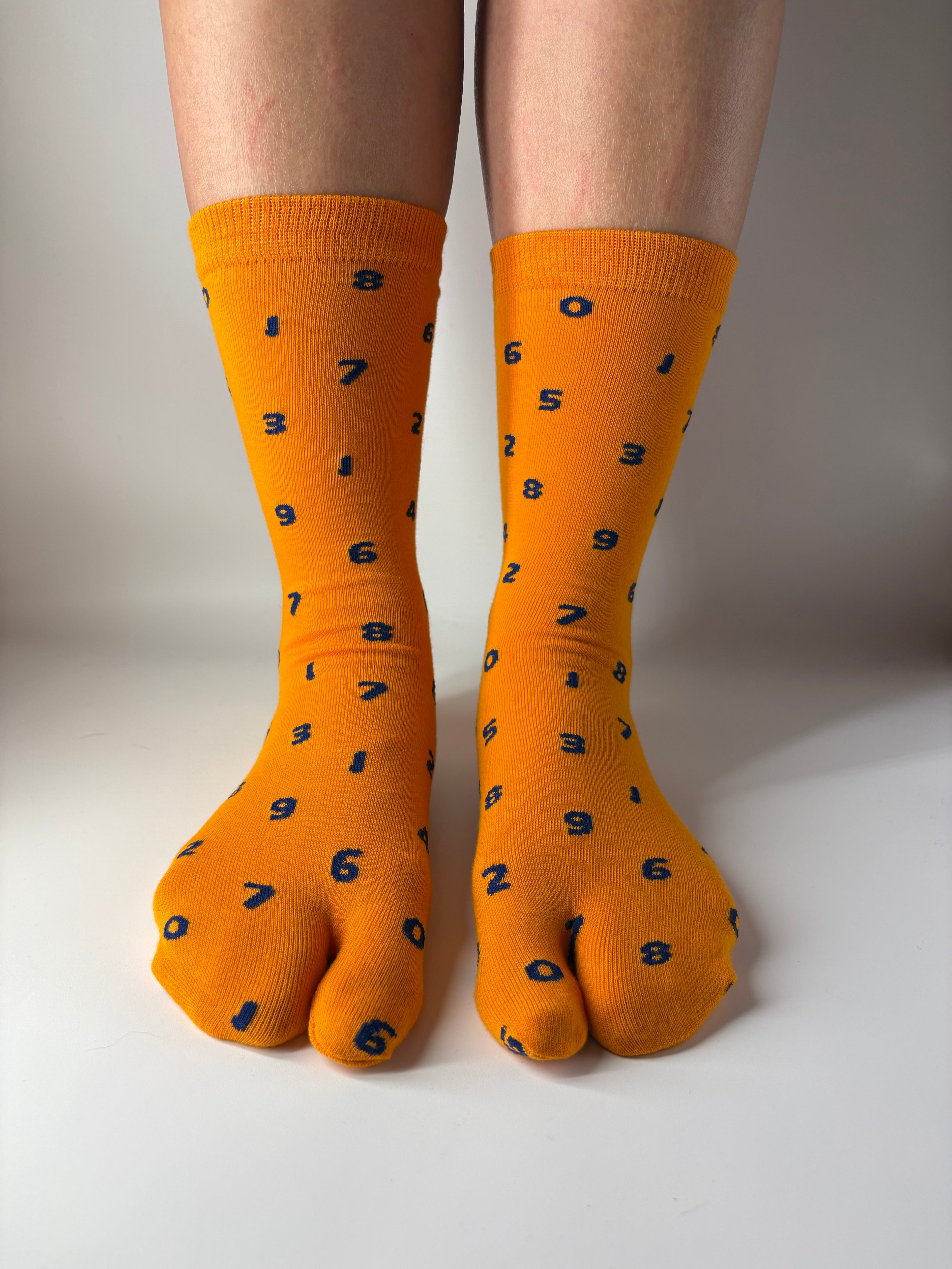 A pair of bright orange crew socks with black printed numbers, worn on feet resting against a plain white wall.