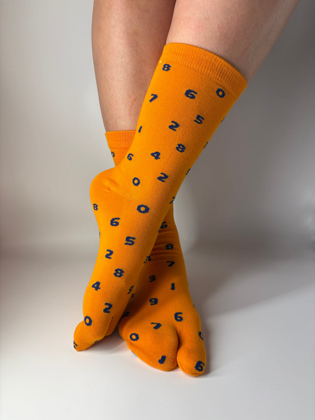 A pair of bright orange tabi socks with small navy blue numbers, worn on a person's feet and crossed over against a plain light gray background.