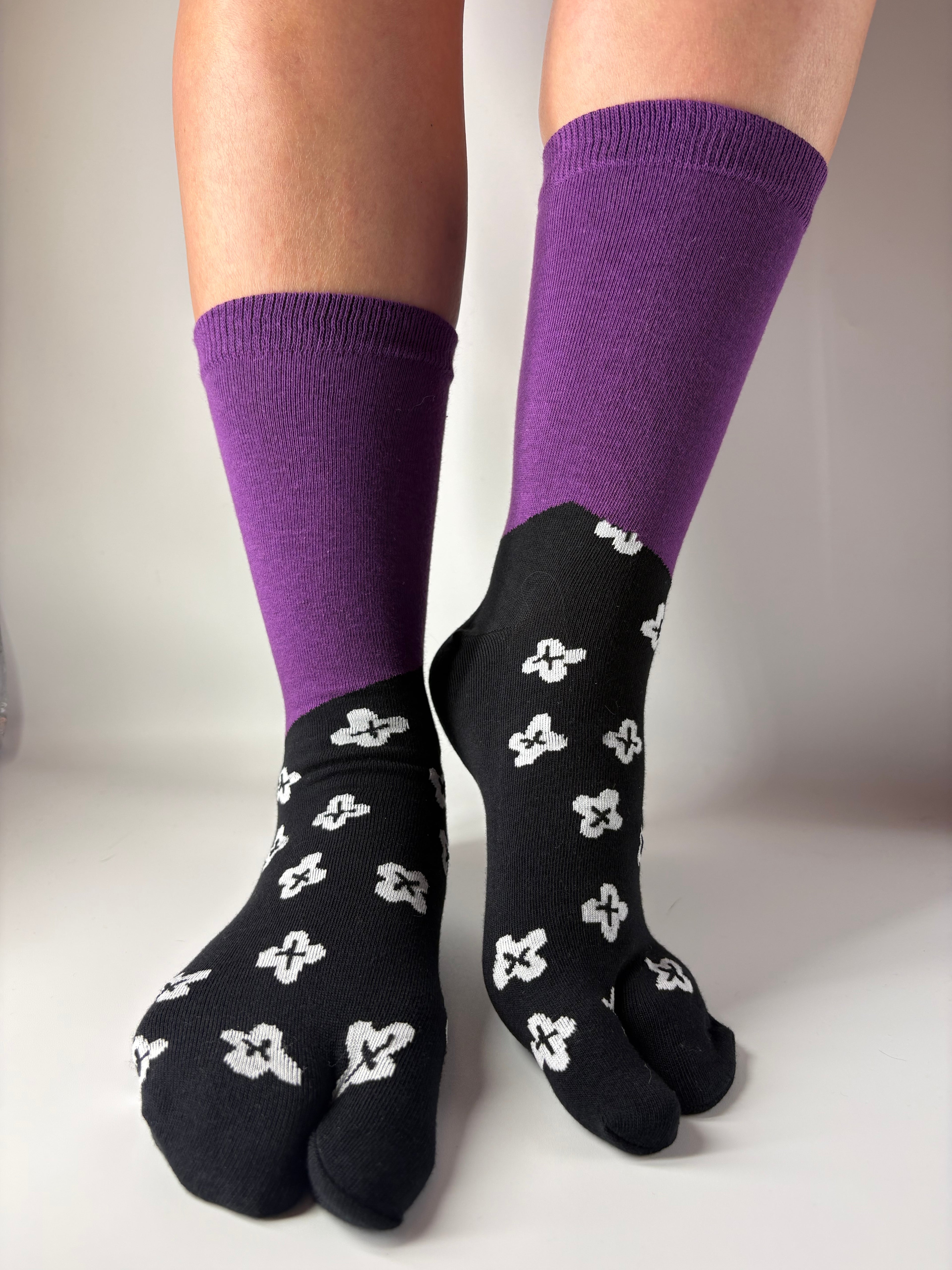 Black tabi socks with white floral patterns and purple cuffs worn by a person on a light grey background