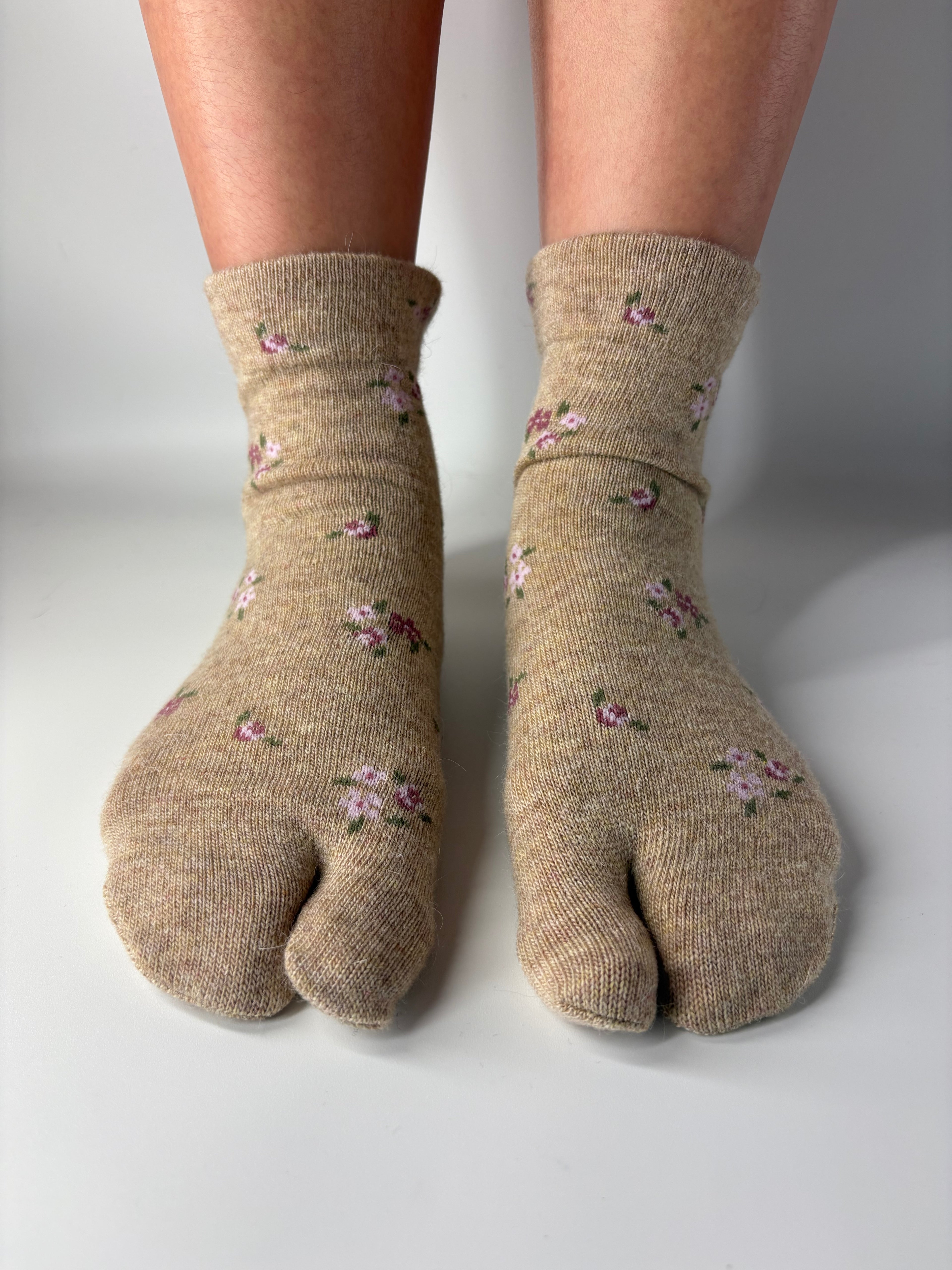 Pair of beige tabi split toe wool socks with floral patterns worn on a white background