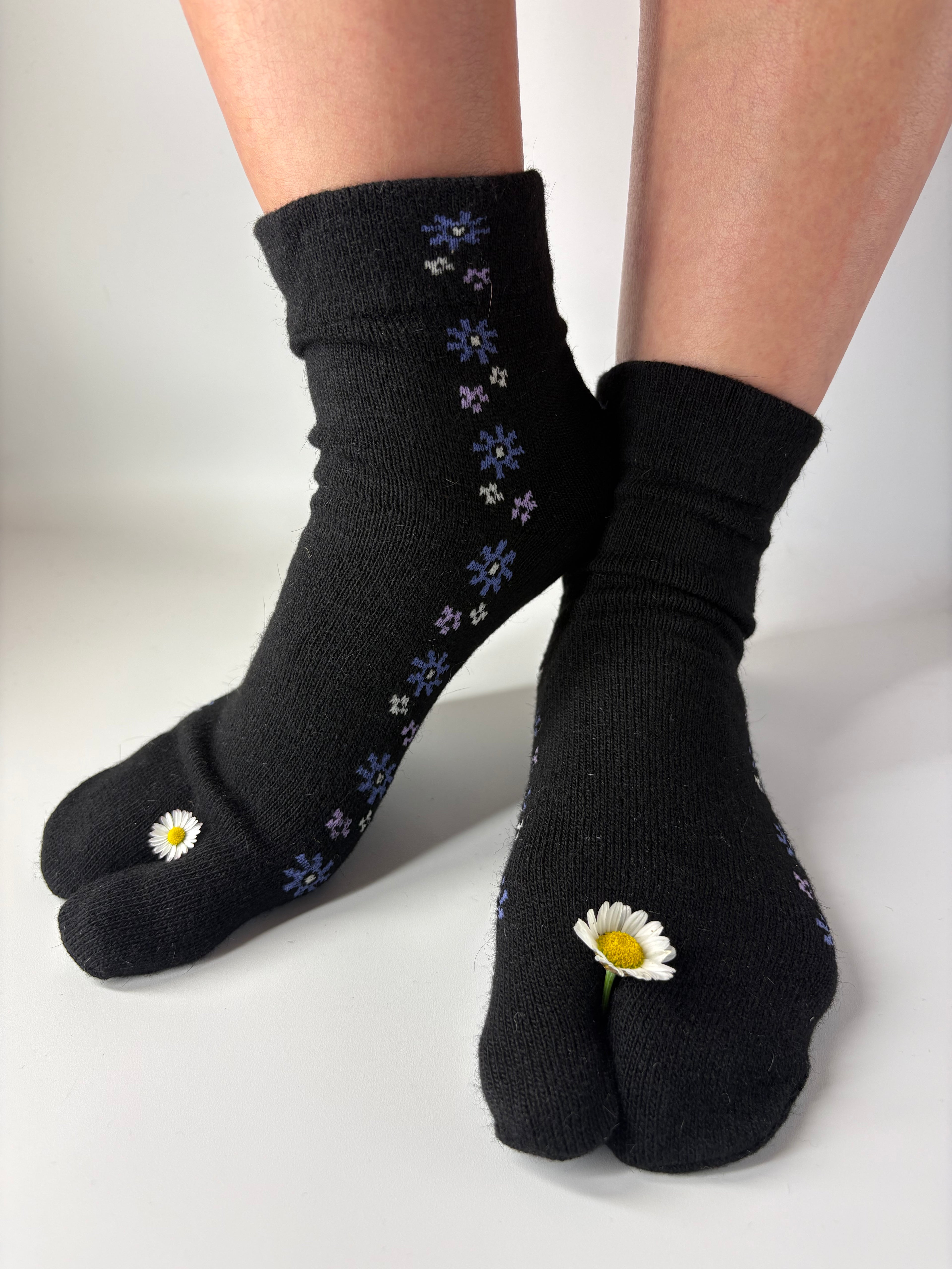 A pair of black tabi socks with small daisy and blue flower patterns, shown on feet against a plain white background.