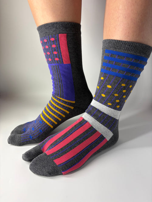 Side view of asymmetrical multi coloured abstract graphic print on grey socks , standing on a white floor with a split toe japanese sock design. Sock is made of cotton natural fibres