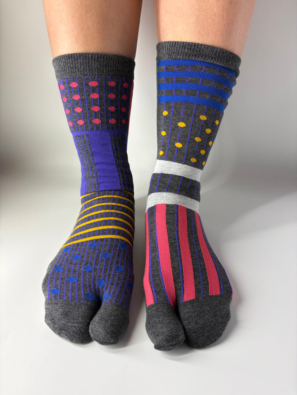 Front view of asymmetrical multi coloured abstract graphic print on grey socks , standing on a white floor with a split toe japanese sock design. Sock is made of cotton natural fibres
