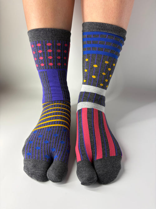 Front view of asymmetrical multi coloured abstract graphic print on grey socks , standing on a white floor with a split toe japanese sock design. Sock is made of cotton natural fibres