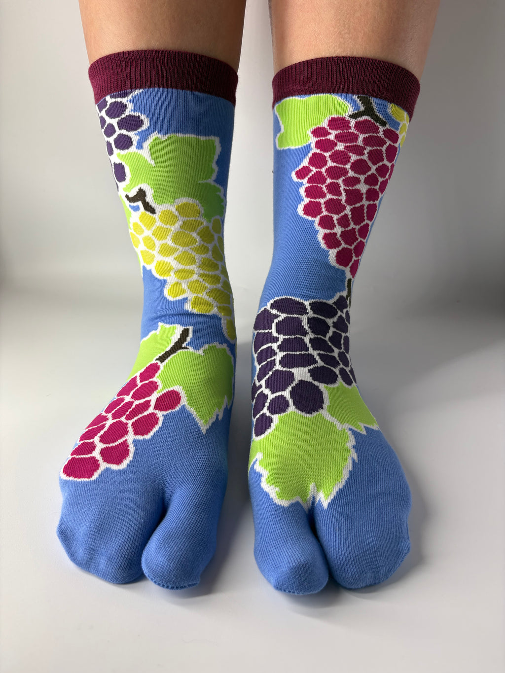 Tabi Co. grape print tabi socks in cornflower blue with burgundy cuff, featuring multicolour grape cluster design in pink, purple and yellow-green, worn on legs against white background