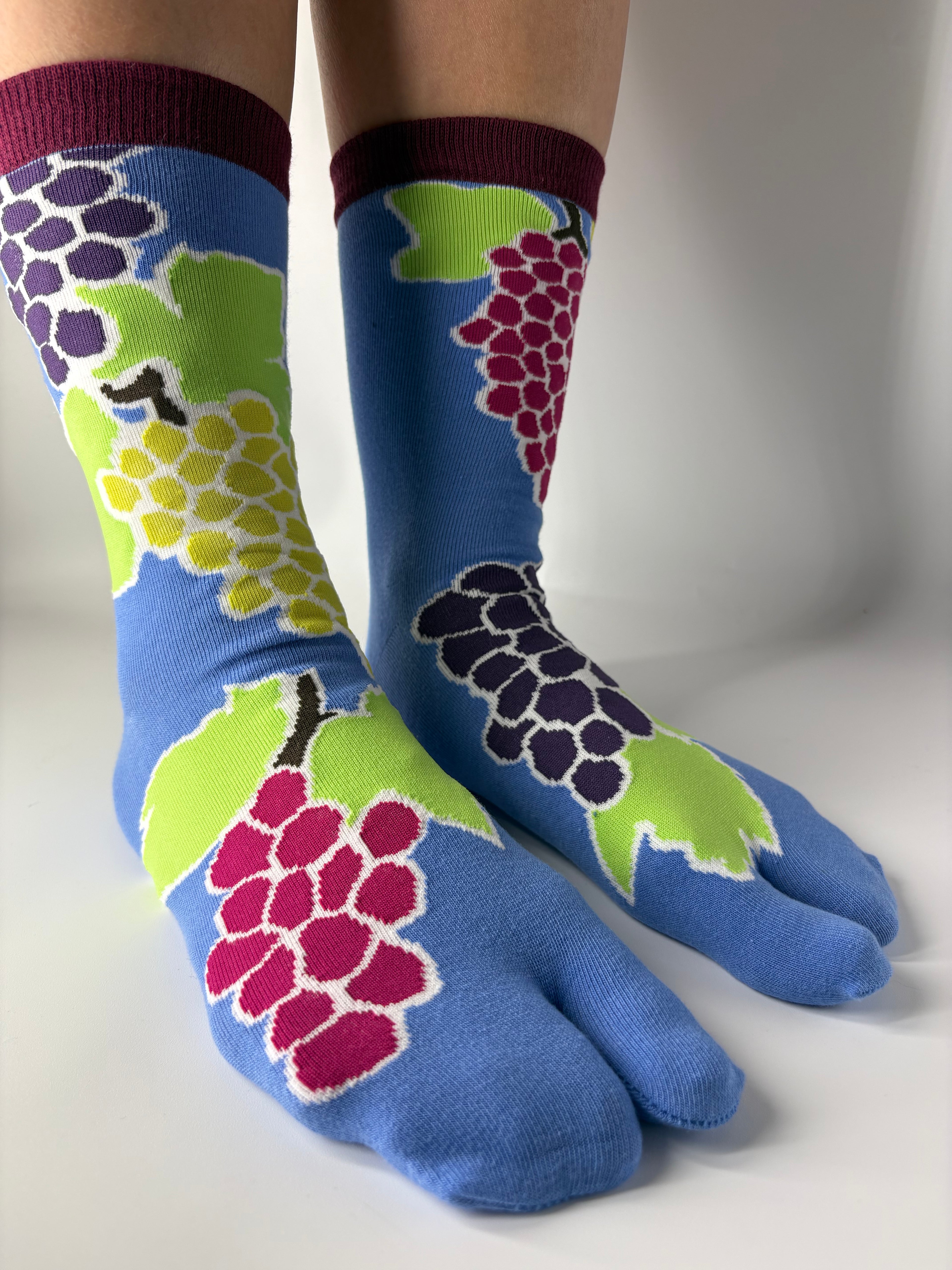 Tabi Co. grape print tabi socks in cornflower blue with burgundy cuff, featuring multicolour grape cluster design in pink, purple and yellow-green, worn on legs against white background