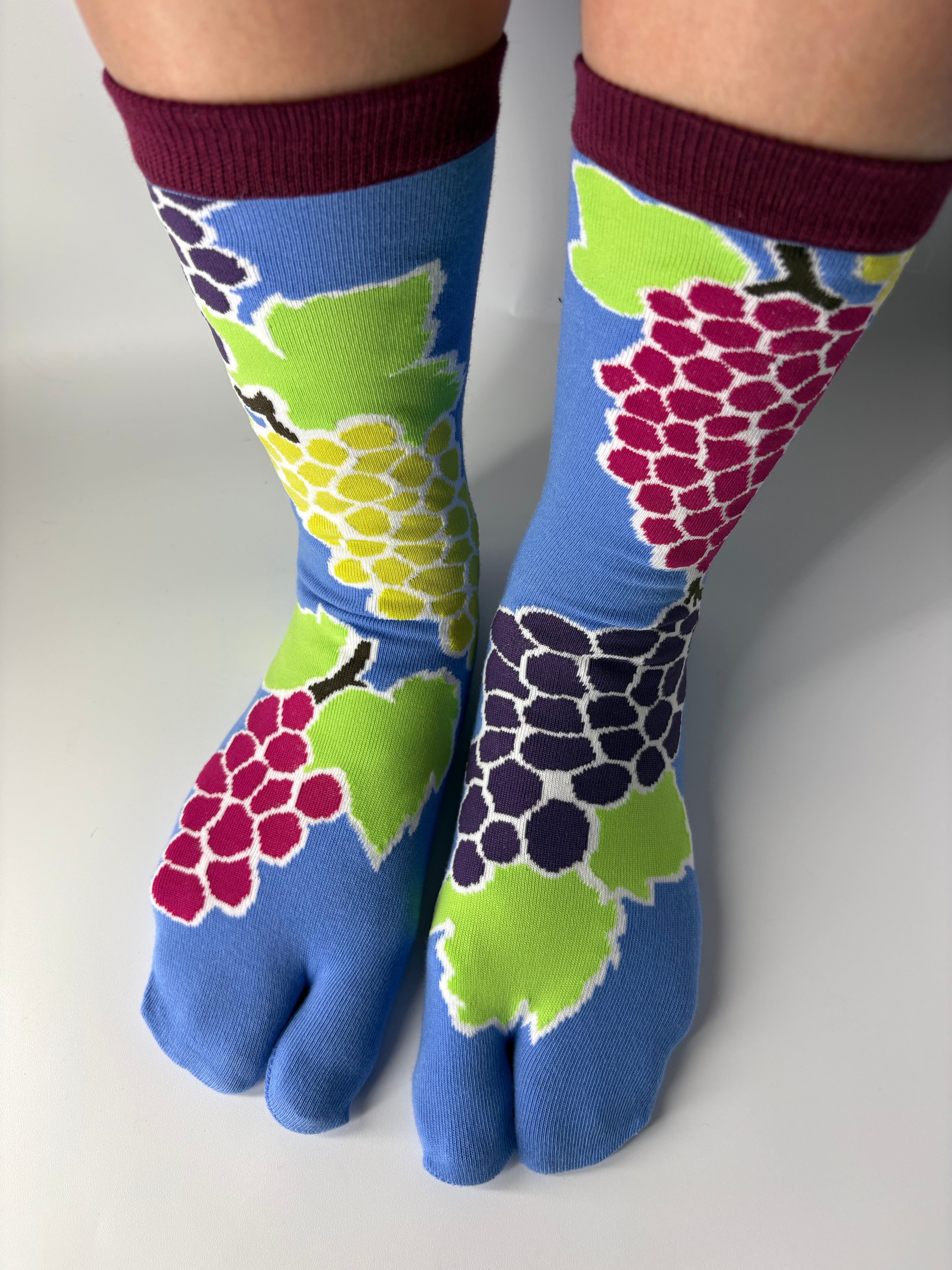 Tabi Co. grape print tabi socks in cornflower blue with burgundy cuff, featuring multicolour grape cluster design in pink, purple and yellow-green, worn on legs against white background