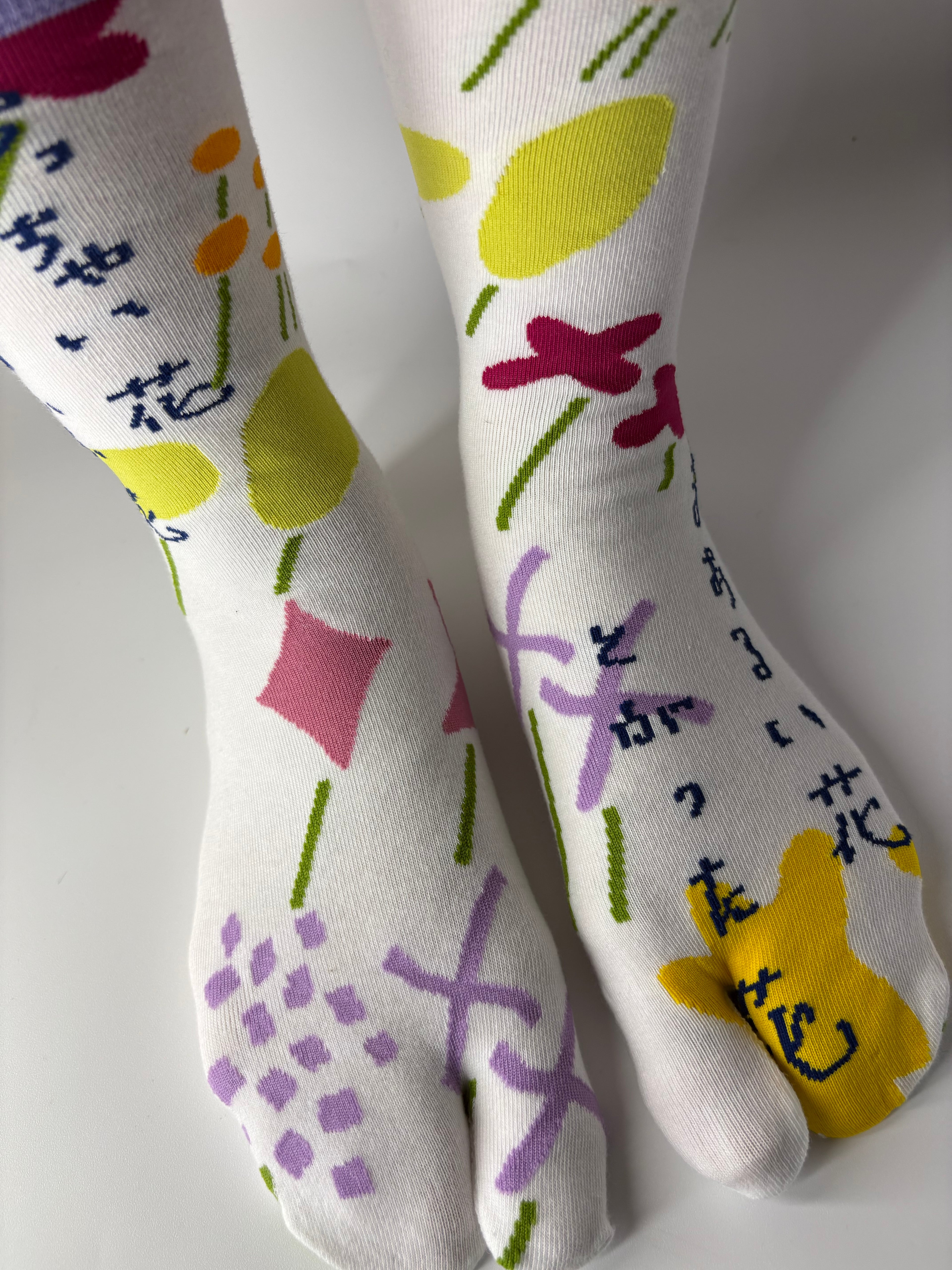 front view of asymmetrical multi coloured abstract graphic print on white socks , standing on a white floor with a split toe japanese sock design. Sock is made of cotton natural fibres