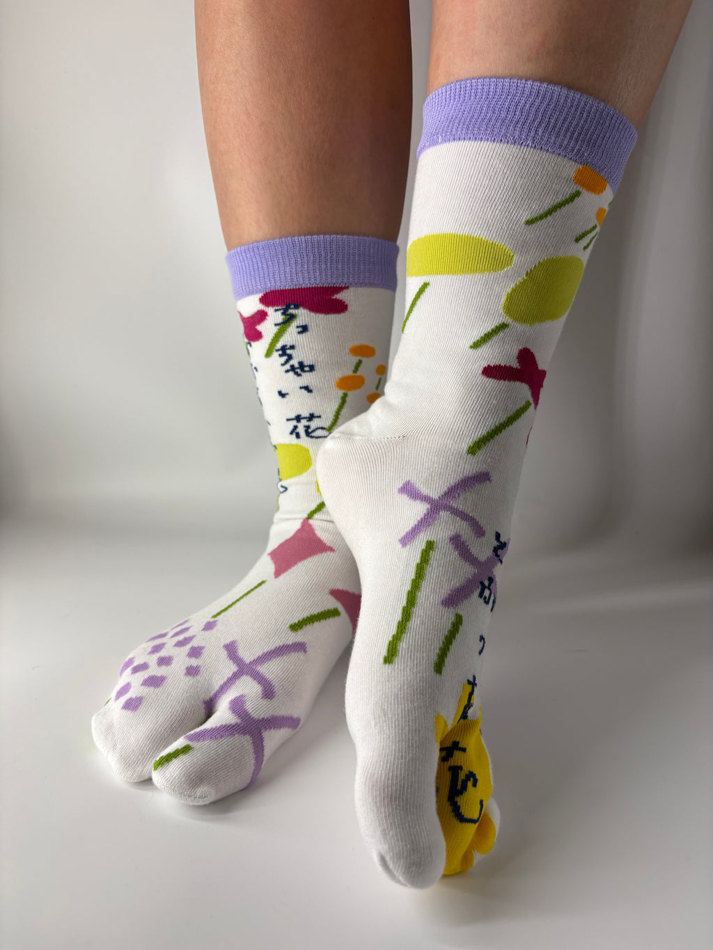 Side view of asymmetrical multi coloured abstract graphic print on white socks , there is a lilac ribbed cuff and model is standing on a white floor with a split toe japanese sock design. Sock is made of cotton natural fibres