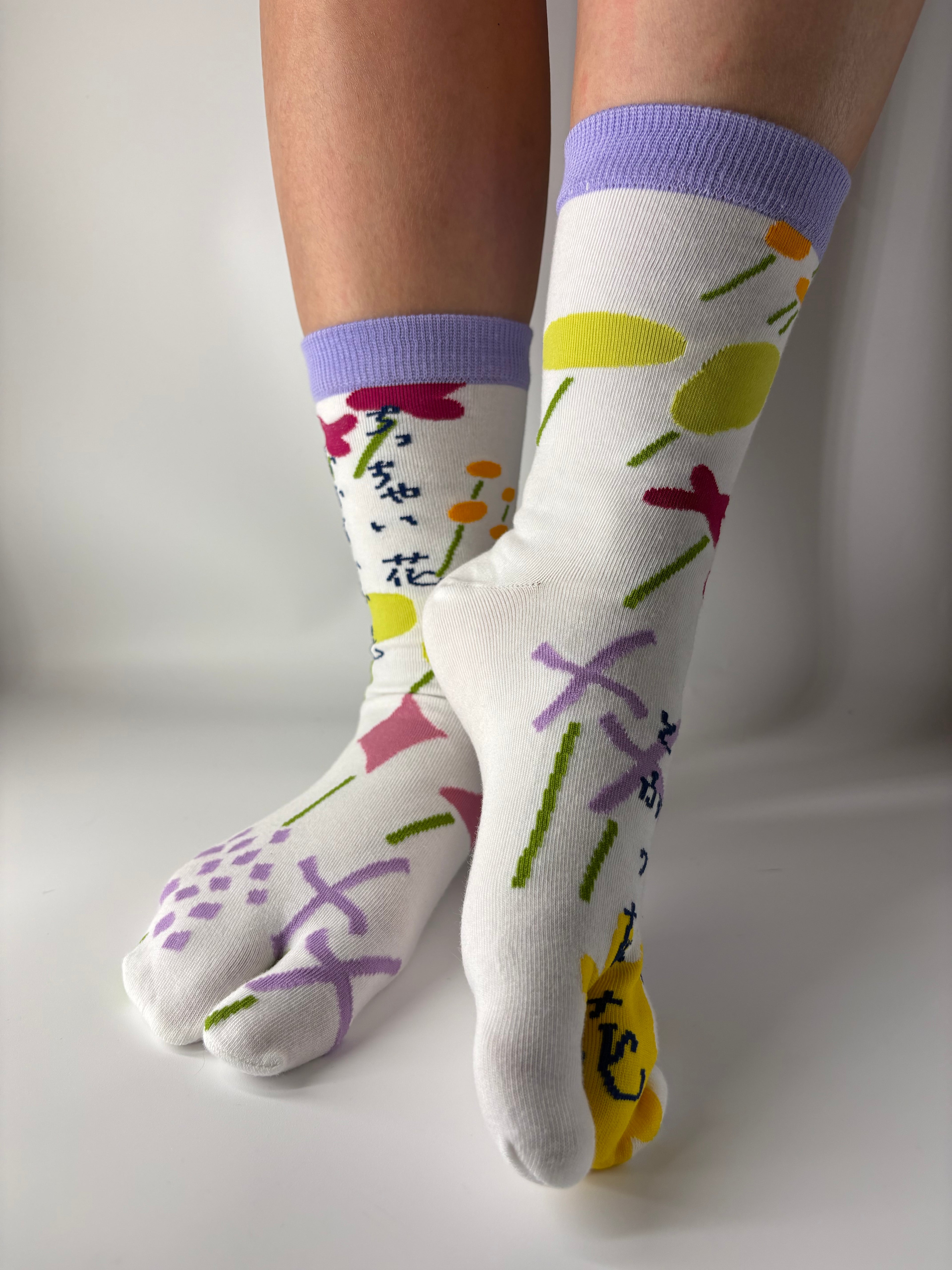 Side view of asymmetrical multi coloured abstract graphic print on white socks , there is a lilac ribbed cuff and model is standing on a white floor with a split toe japanese sock design. Sock is made of cotton natural fibres