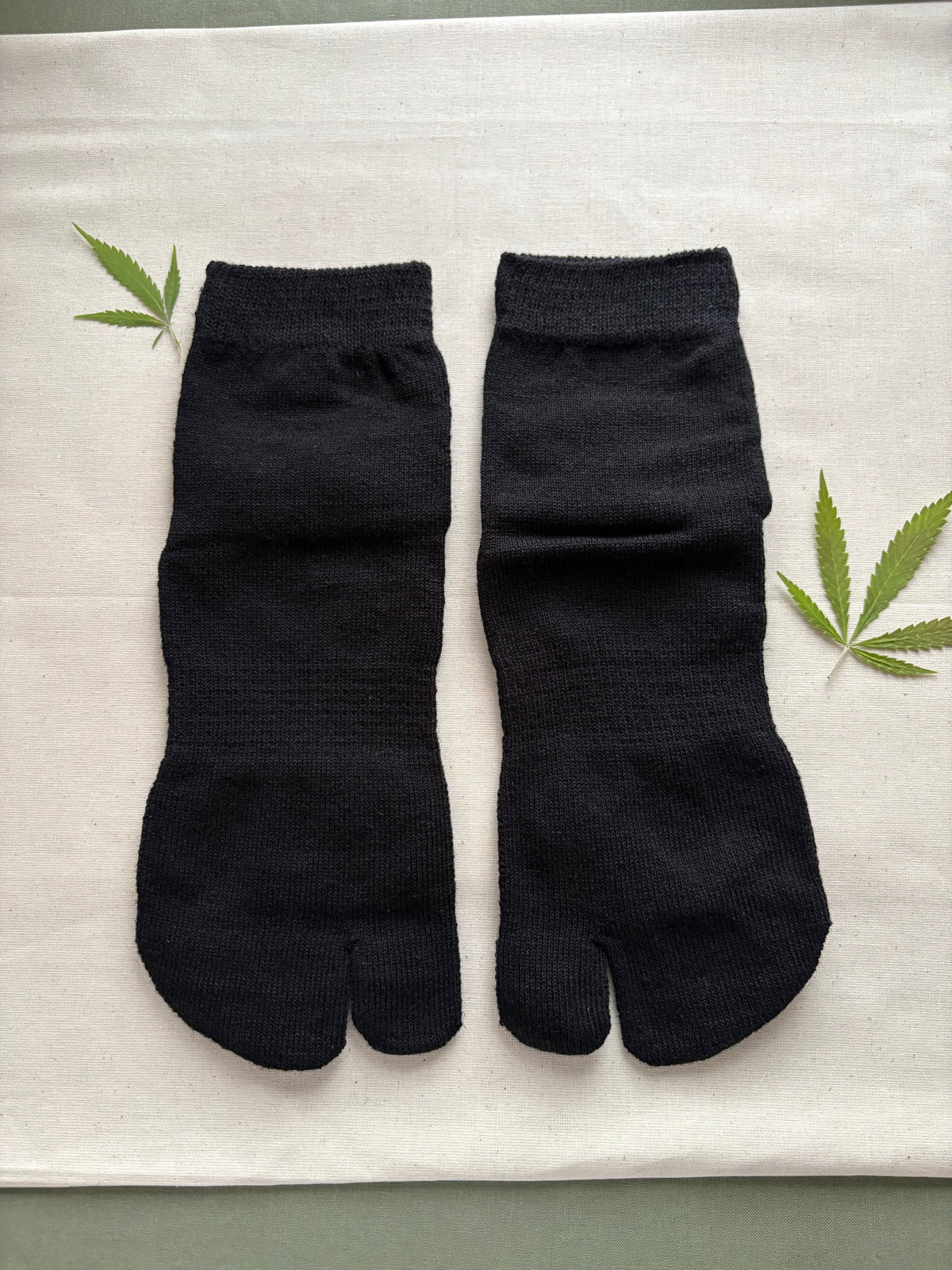 A pair of black ribbed tab socks laid flat on a light beige fabric surface, with two green hemp leaves placed near the edges as props.