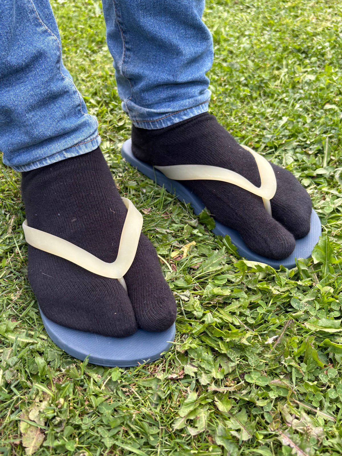 A person wearing blue jeans, black socks, and split-toe flip-flop sandals with cream straps standing on green grass.