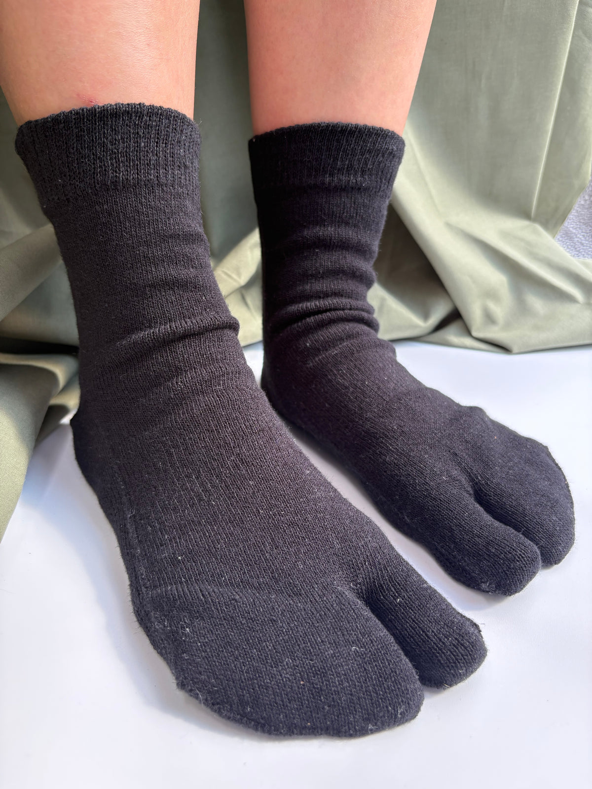 A person wearing black split toe socks, shown from the calves down against a light background with a sage green fabric backdrop.