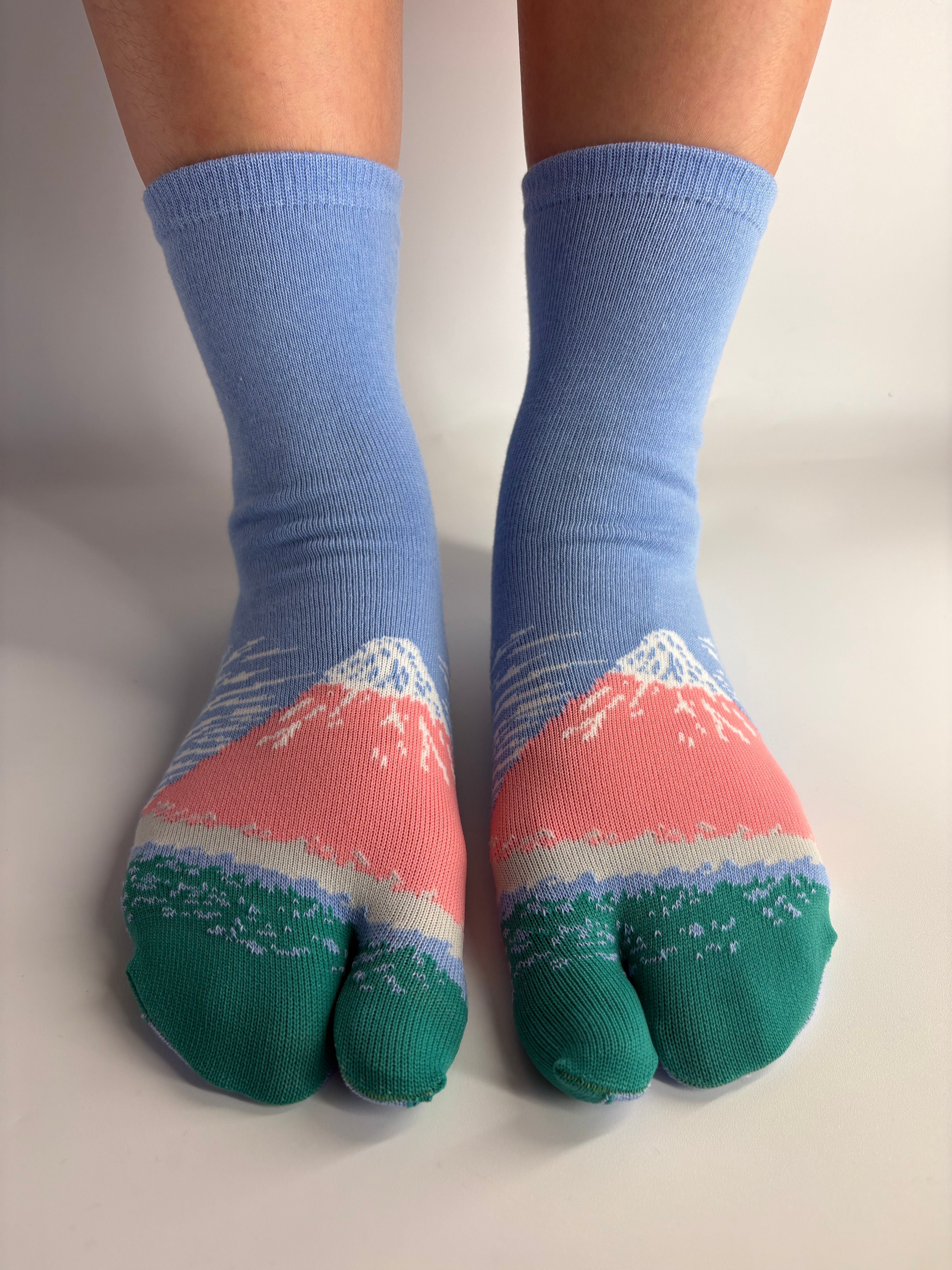 A pair of split-toe socks worn on feet, featuring a light blue ribbed leg section, a coral pink and white mountain graphic, and a green toe cap with a white dotted pattern.