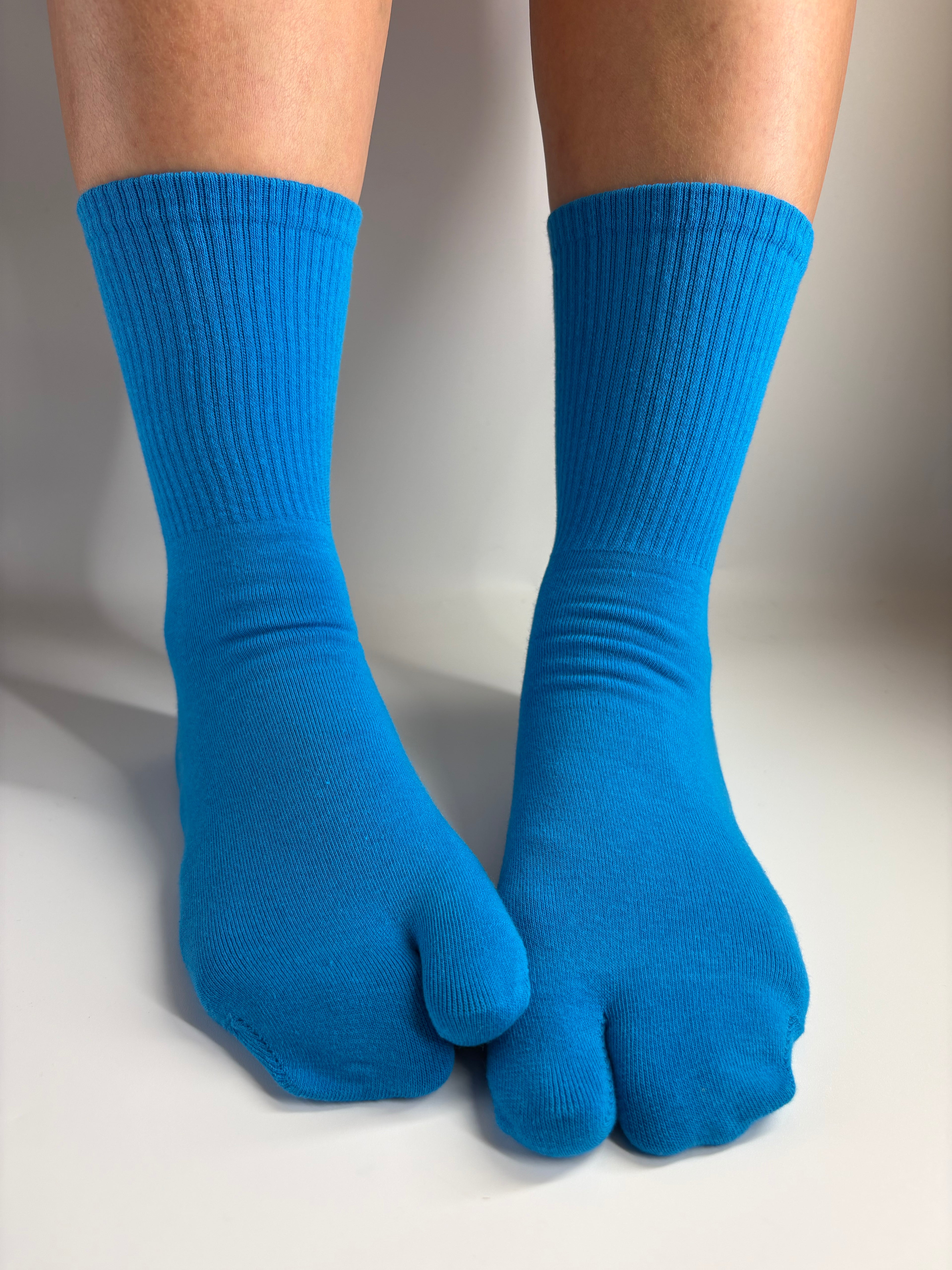 Bright blue ribbed tabi split toe socks worn on a person's feet, shown against a plain light gray studio background.