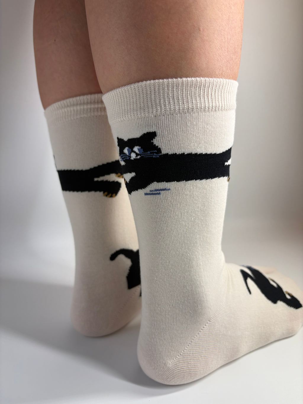 A close-up of a person's legs wearing off-white crew socks with black cat silhouettes and a small black-and-white cat graphic near the cuff.