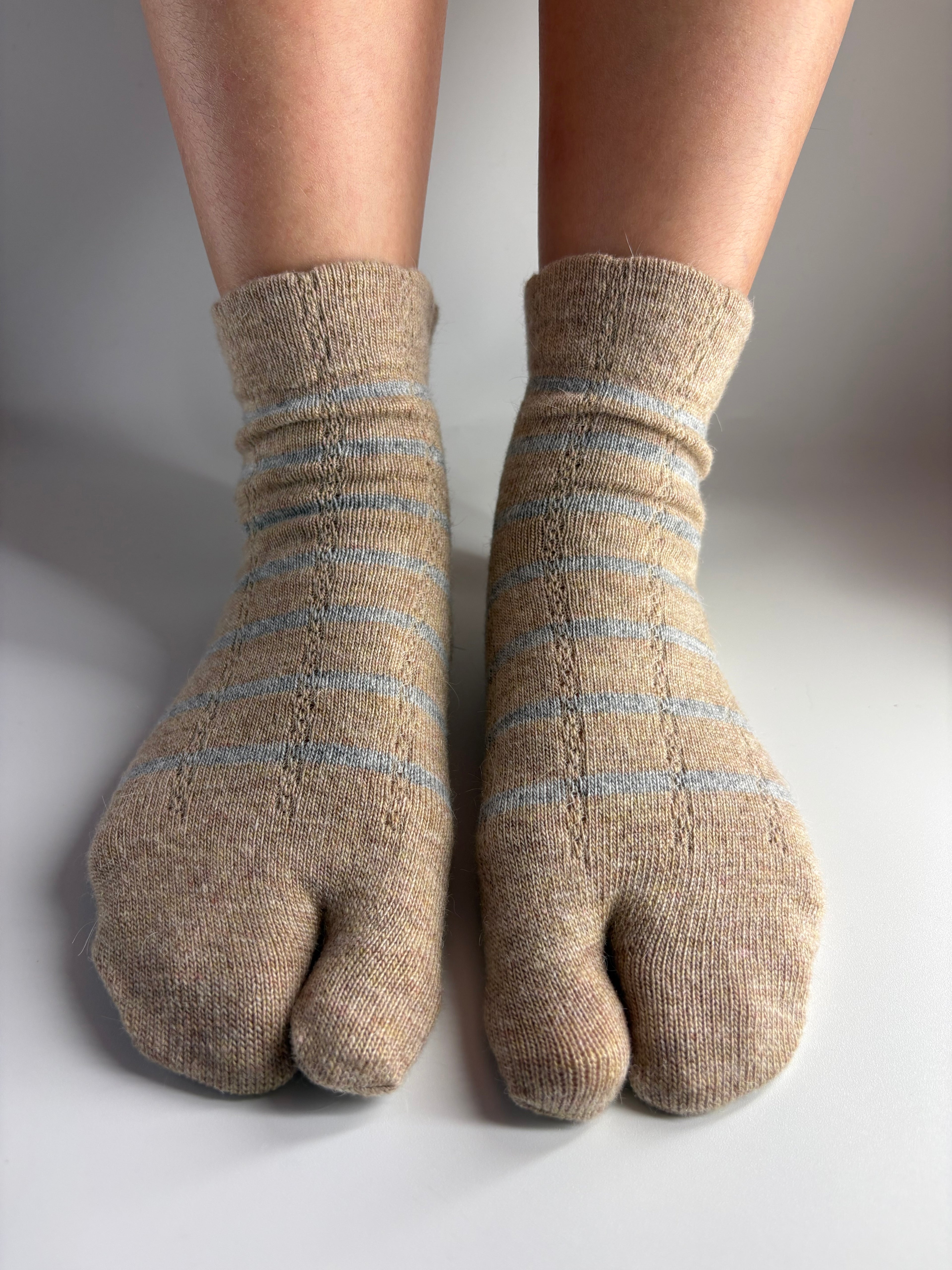 A pair of beige knit tabi socks with light grey stripes worn on feet, shown from a top-down view on a light gray background.