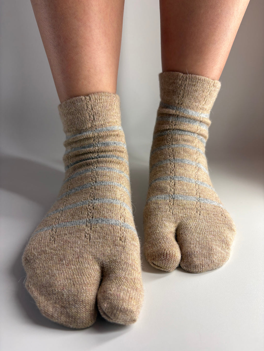 A pair of beige split-toe tabi foot socks with light grey stripes, shown worn on feet against a plain light gray background.