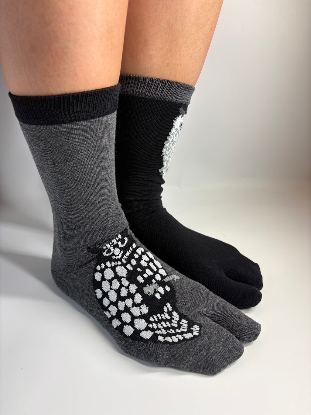 A pair of split-toe socks shown on feet, with one sock in heather gray featuring a white owl print and one sock in black featuring a white owl design.