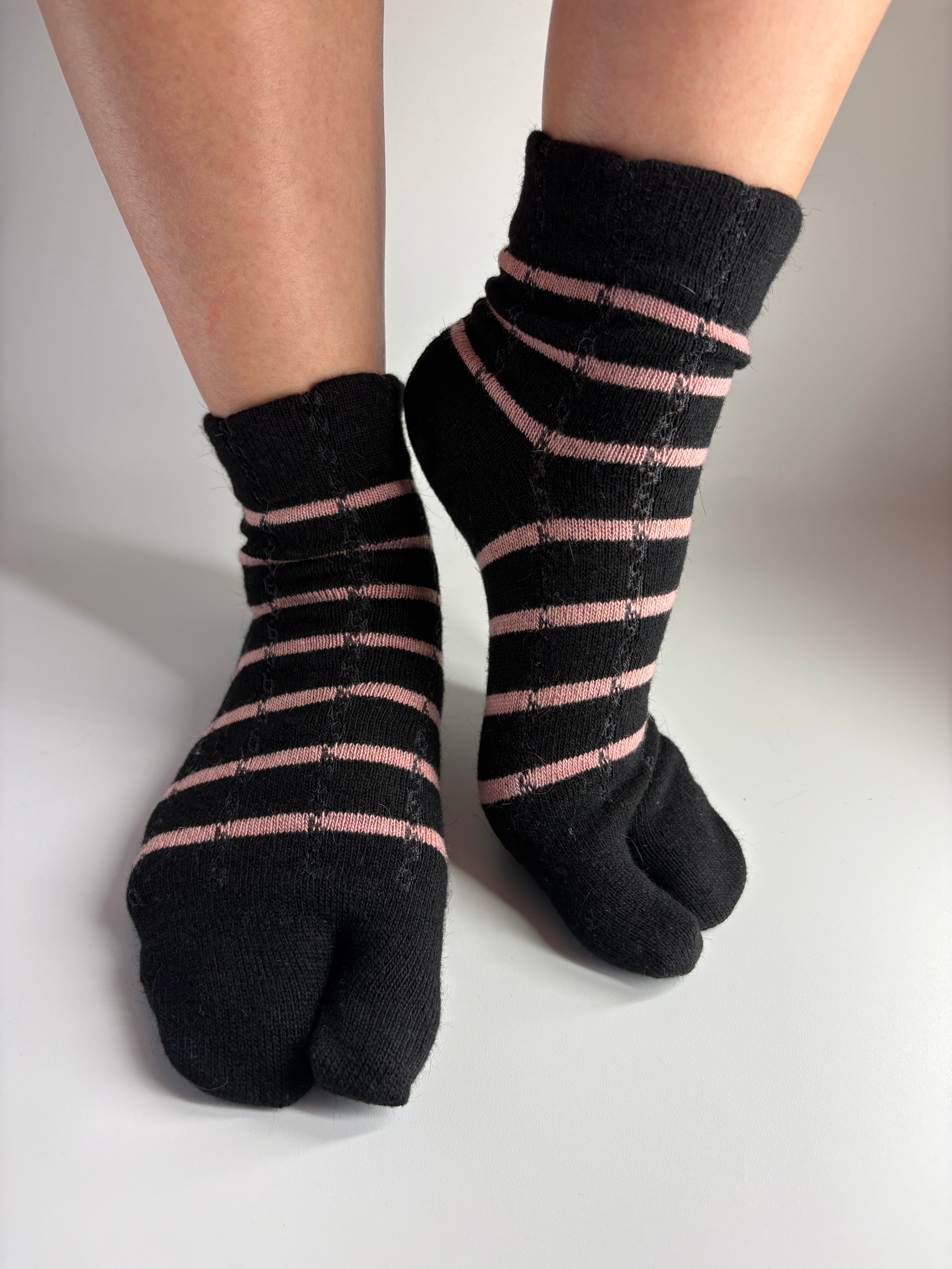 A pair of black tabi socks with thin pink stripes, worn on feet against a plain light gray background.