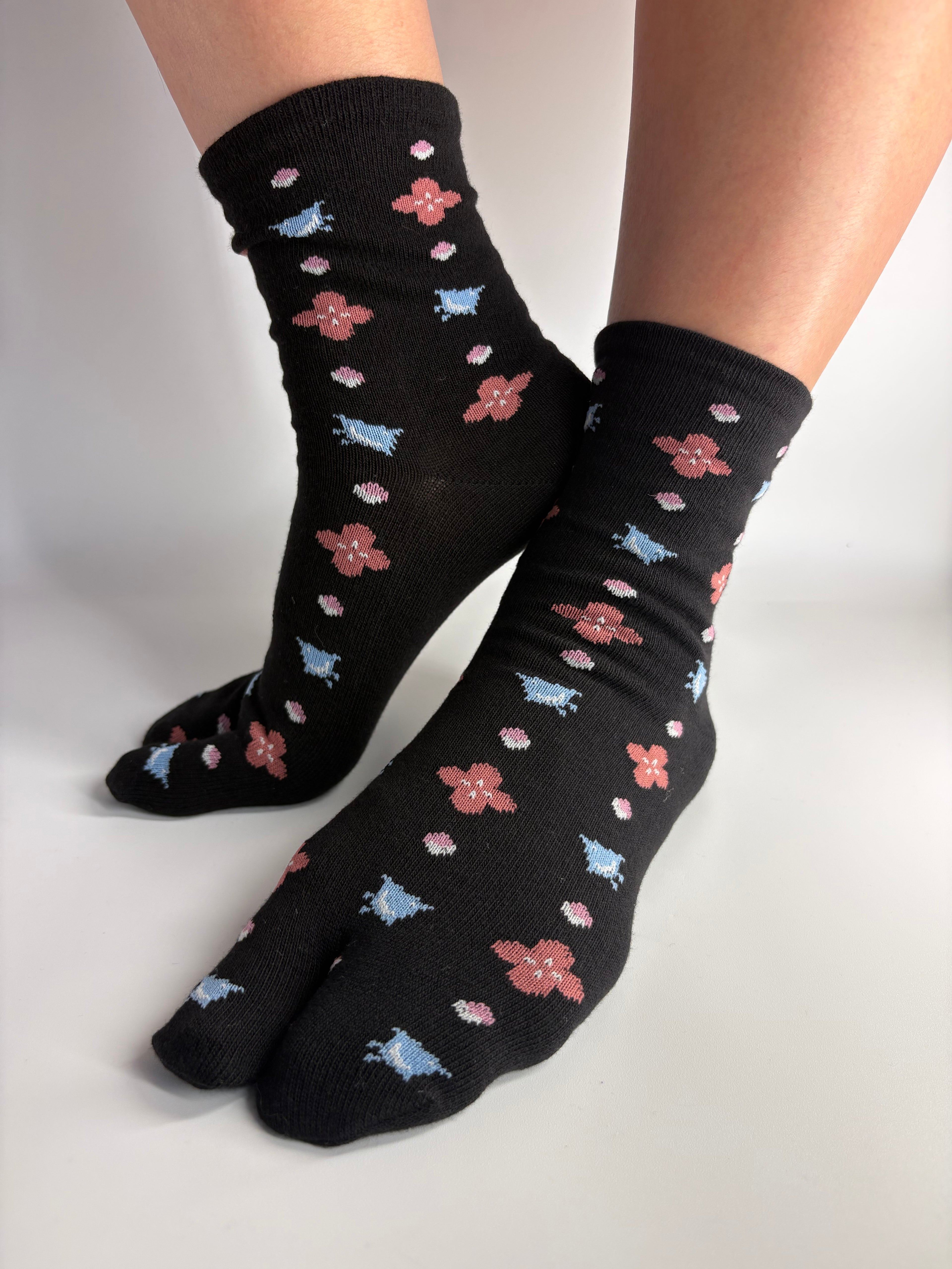 Black crew socks with a repeating pattern of small pink flowers, light blue birds, and white dots, shown worn on a person's feet against a plain white background.