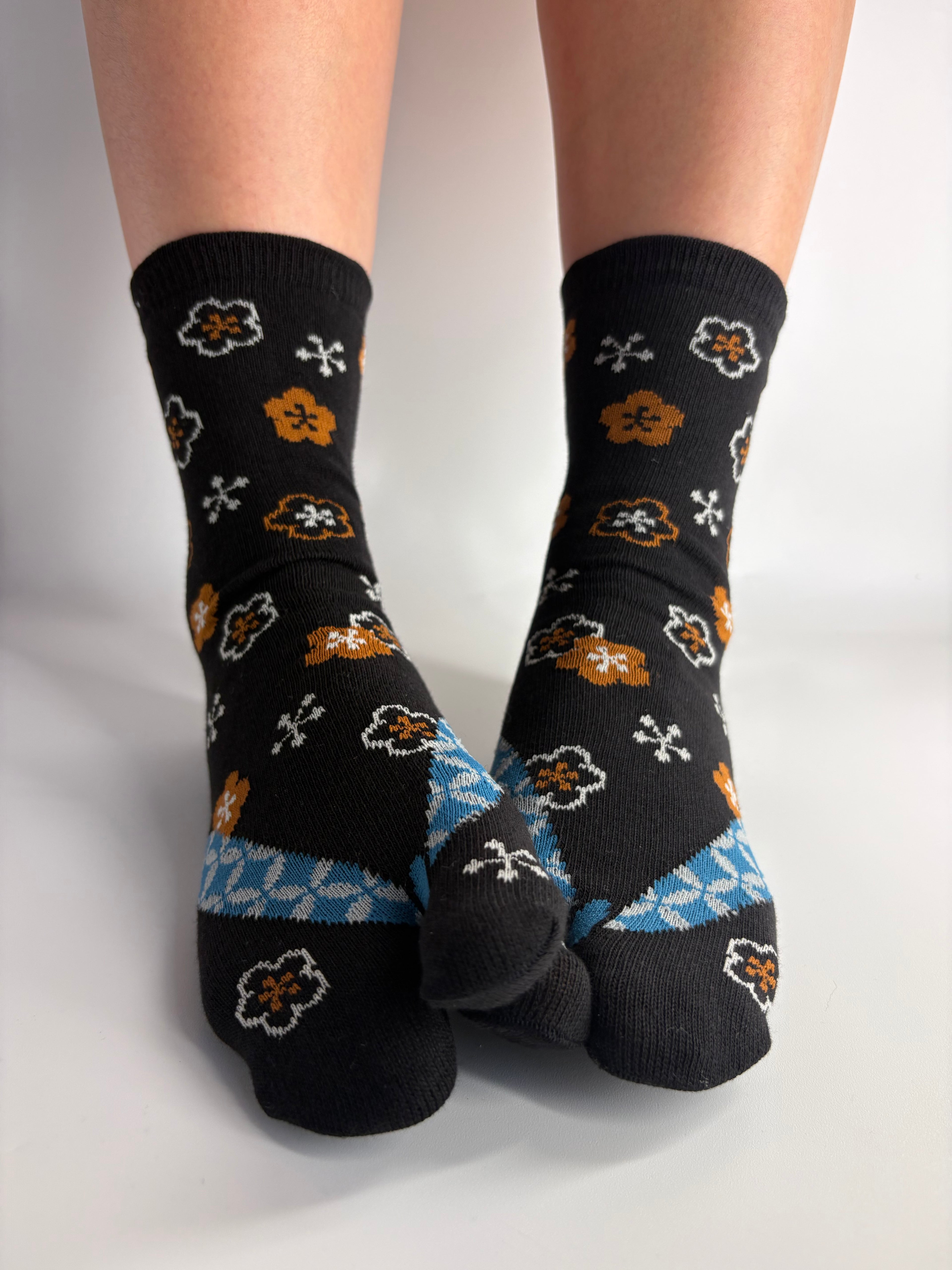 A pair of black crew socks with a pattern of brown floral print, snowflakes and blue and white geometric stripes, worn on feet against a plain white background.