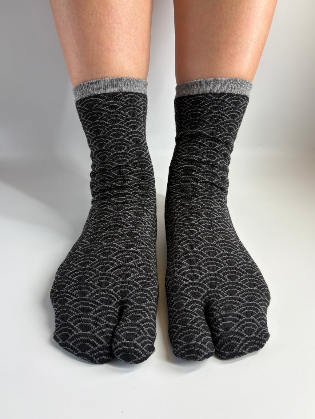 A pair of dark grey tabi socks with a light gray wave pattern of the japanese seigeiha print and ribbed gray cuffs, shown on a person's feet against a light gray background.