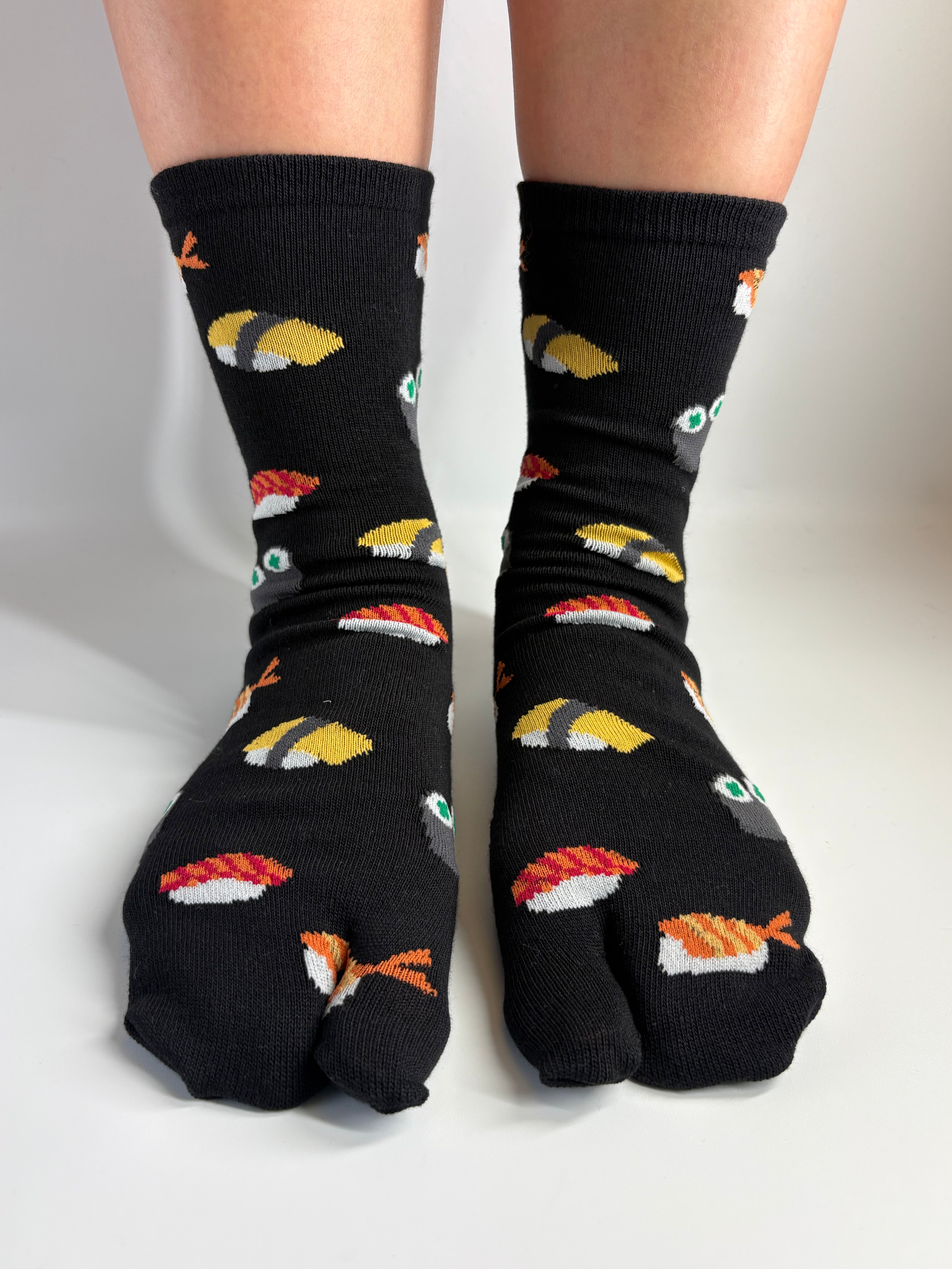 A pair of black split-toe socks with a colorful sushi pattern, worn on feet against a plain white background.