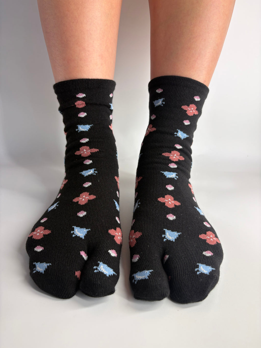 Black tabi socks with a repeating pattern of small pink, light blue birds, and white clover-like flowers, shown worn on feet against a plain light background.