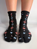 Black tabi socks with a repeating pattern of small pink, light blue birds, and white clover-like flowers, shown worn on feet against a plain light background.