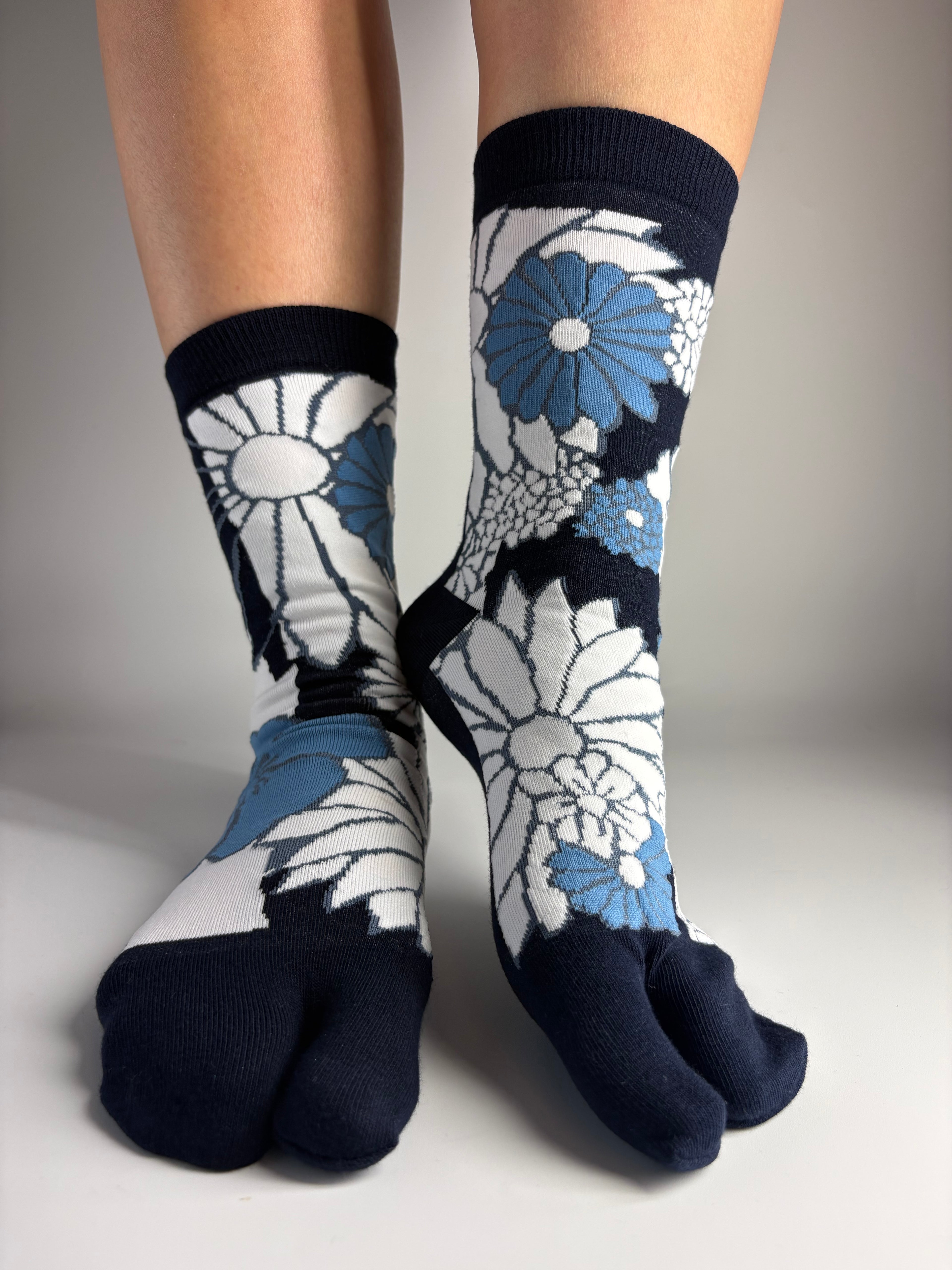 A pair of crew socks with a navy blue toe, heel, and ribbed cuffs, featuring a white and light blue floral dpattern on the sides, shown worn on feet against a neutral gray background.