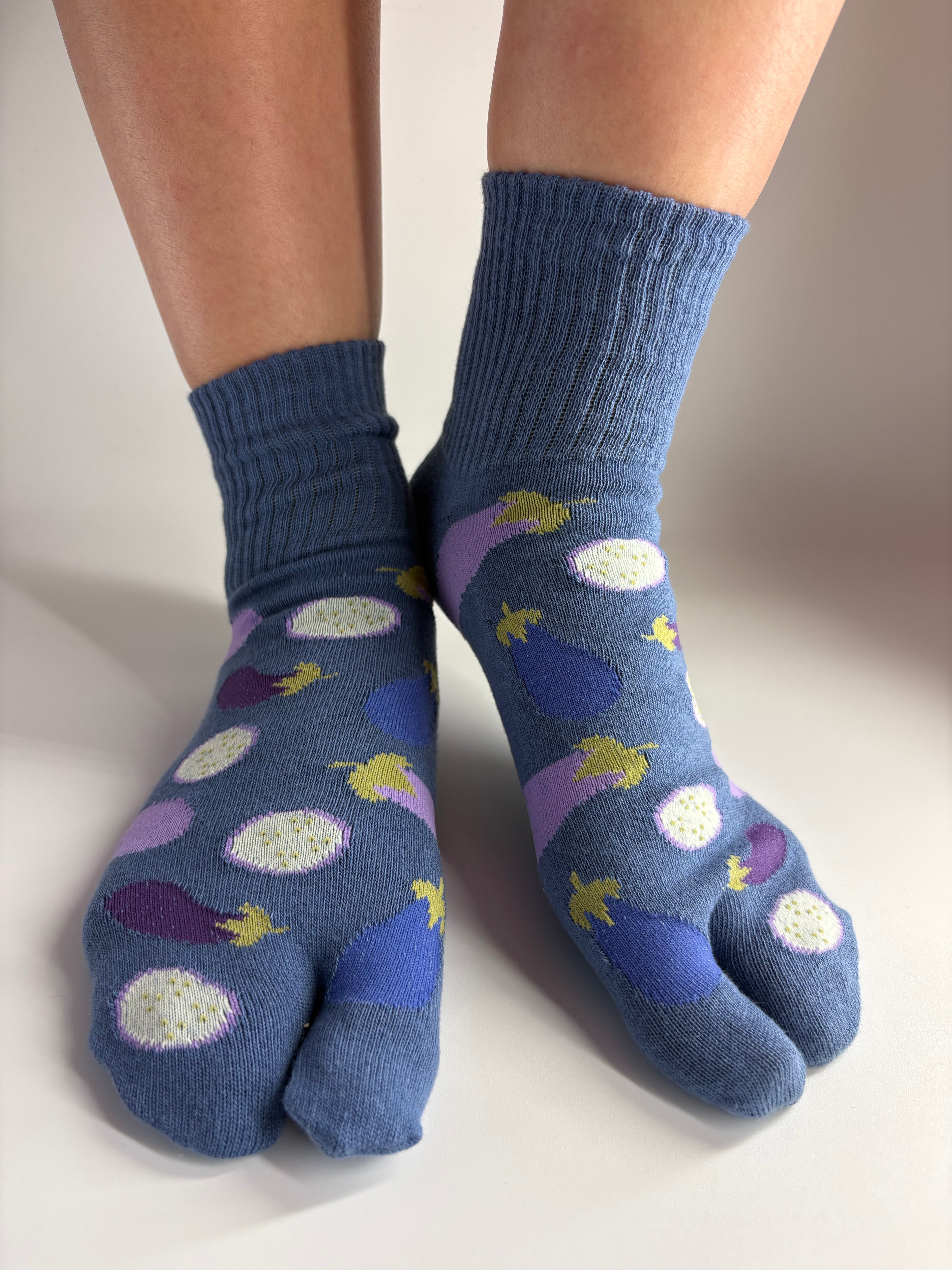 A pair of blue tabi socks with a pattern of purple eggplants, shown worn on feet against a plain light background.