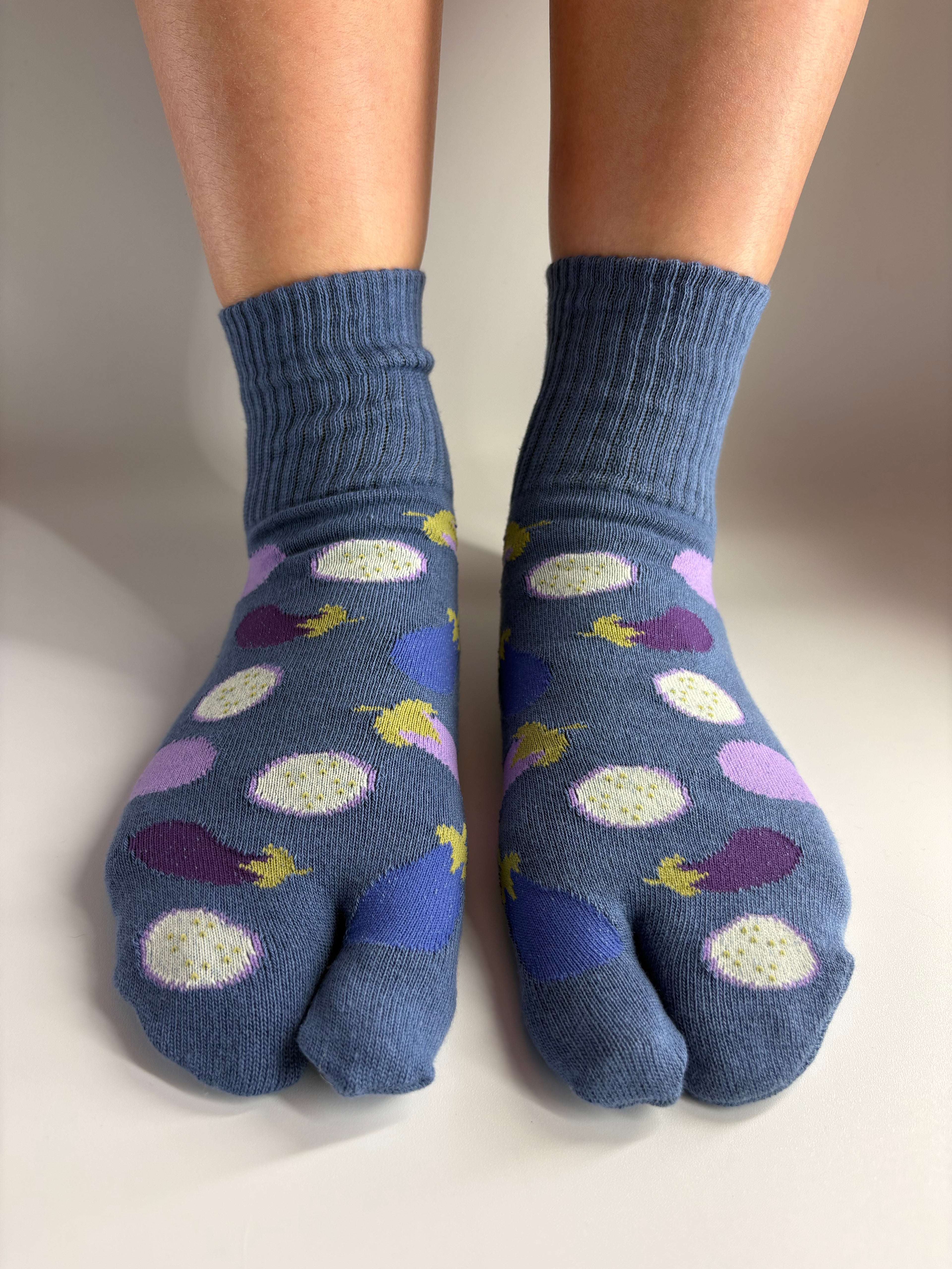 A pair of blue-gray tab socks with a pattern of purple eggplants, shown on feet against a plain light background.
