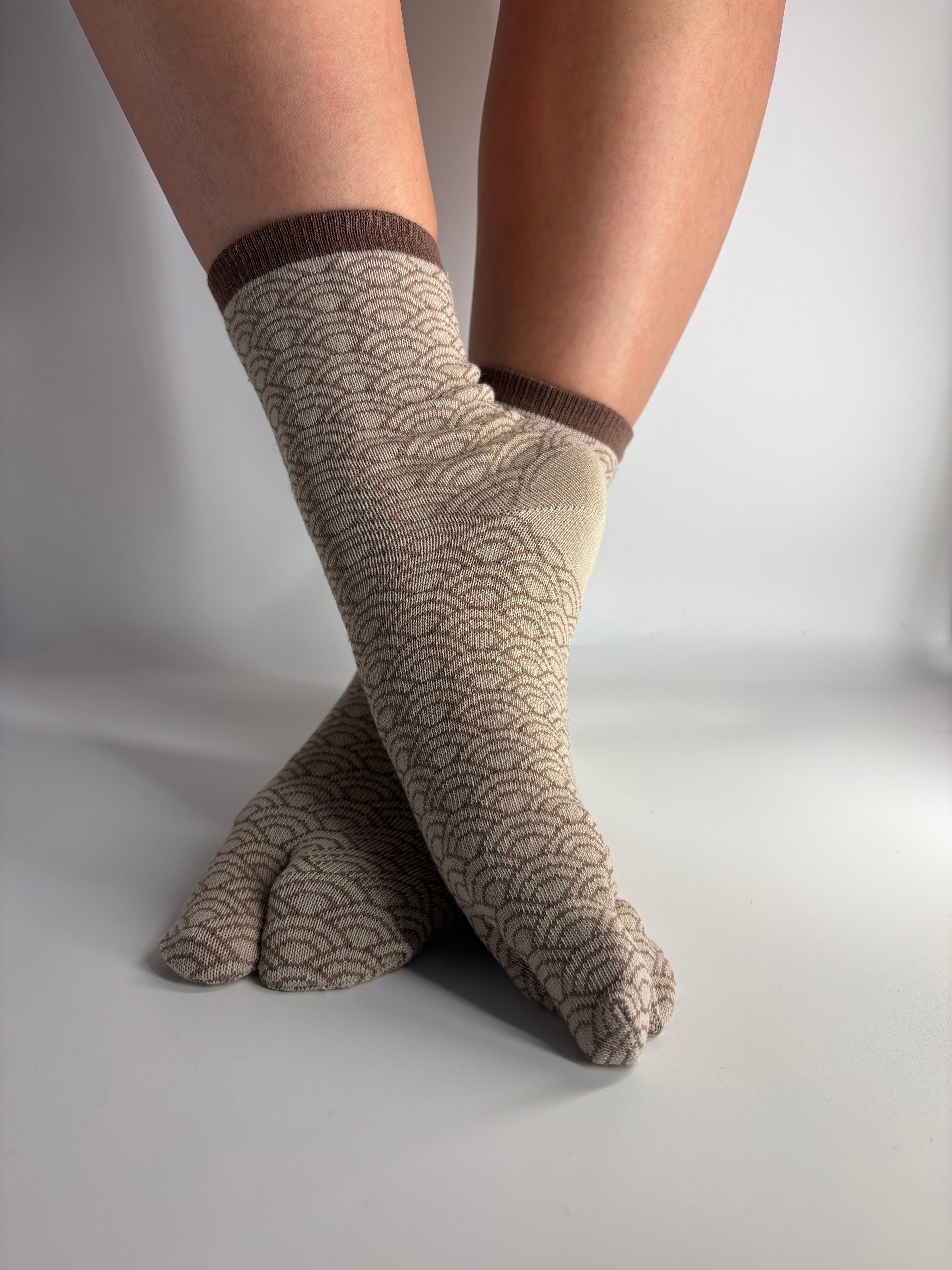 A pair of light beige tabi socks with a traditional japanese pattern and brown ribbed cuffs, shown on feet against a plain white background.