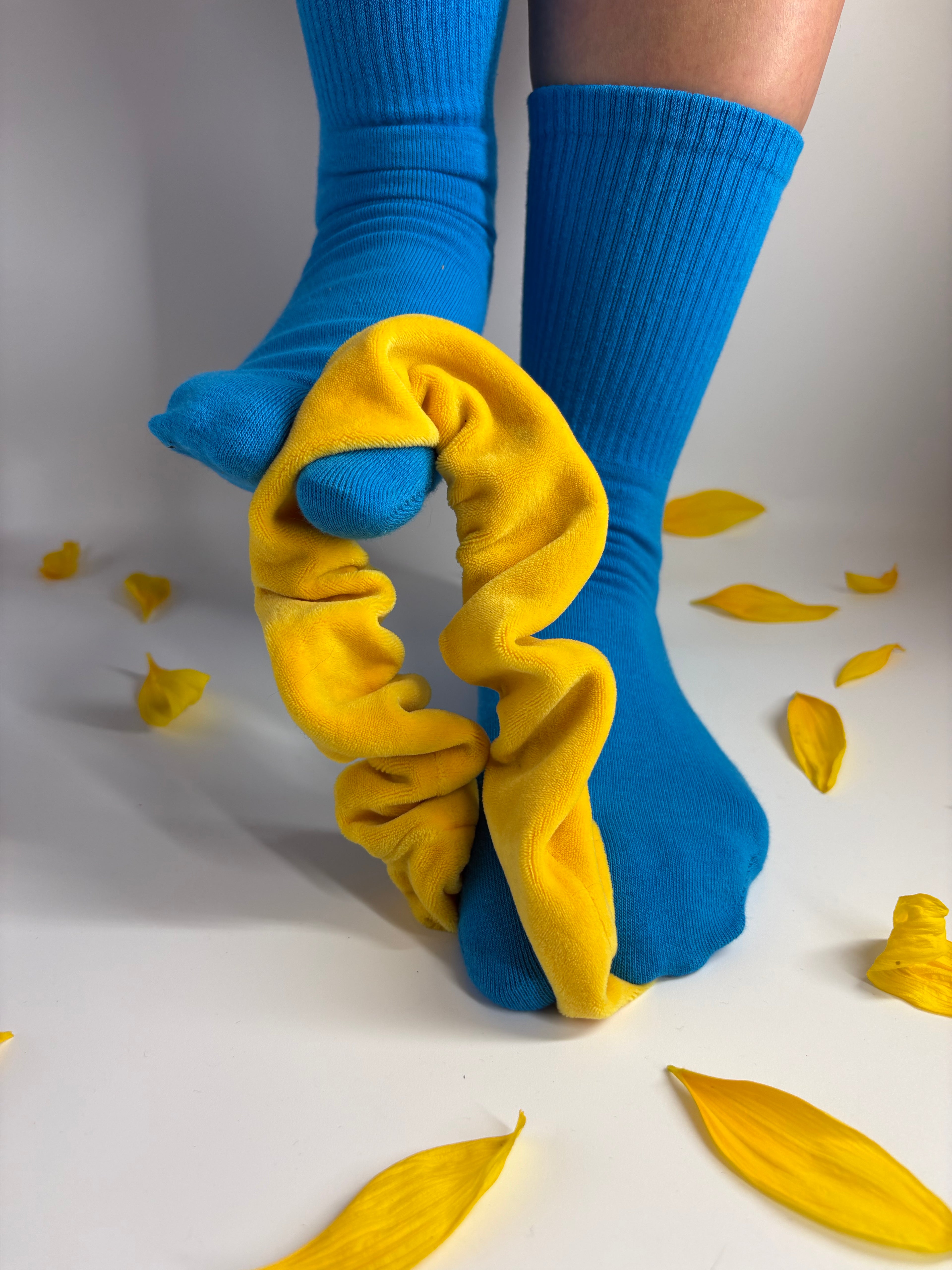 A pair of bright blue crew socks with yellow scrunchie, worn on feet against a white background with scattered yellow flower petals.
