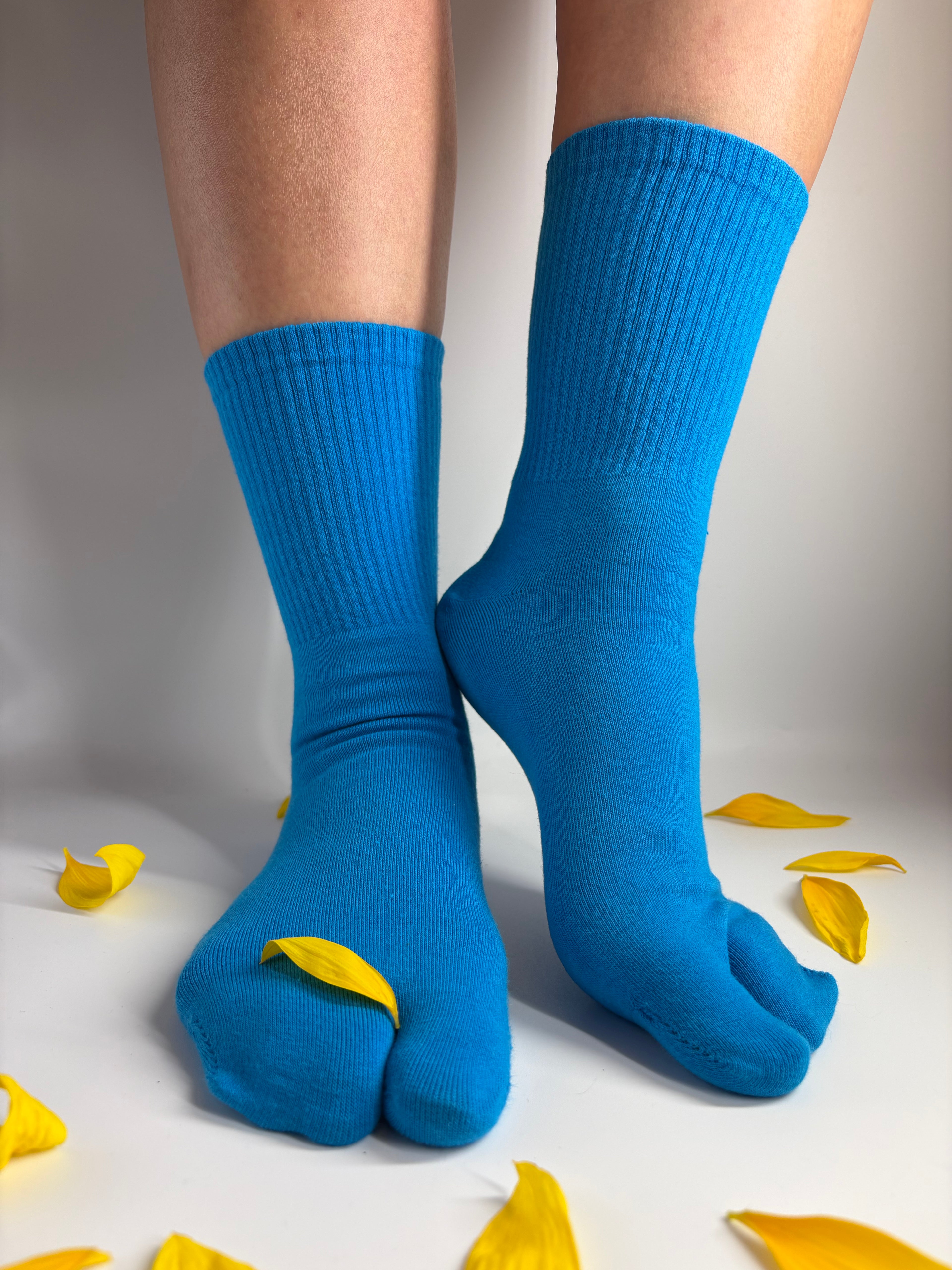 A pair of bright blue ribbed toe socks worn on feet, shown against a white background with scattered yellow flower petals.