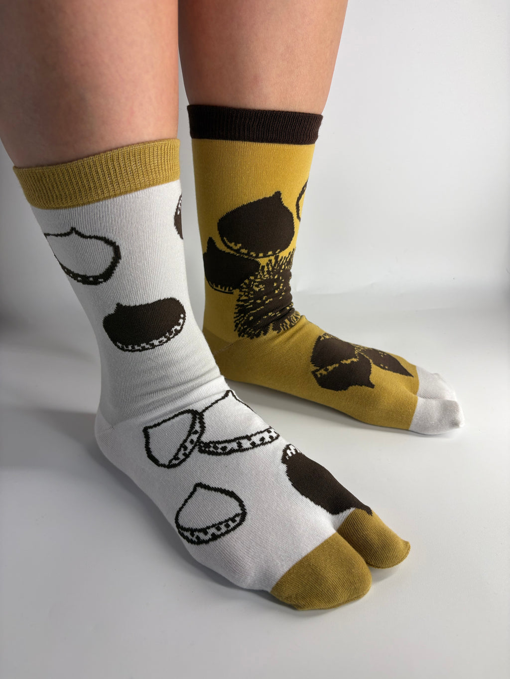 A pair of split-design socks worn on feet, with one sock being white and the other mustard yellow, both featuring dark brown achestnut illustration.