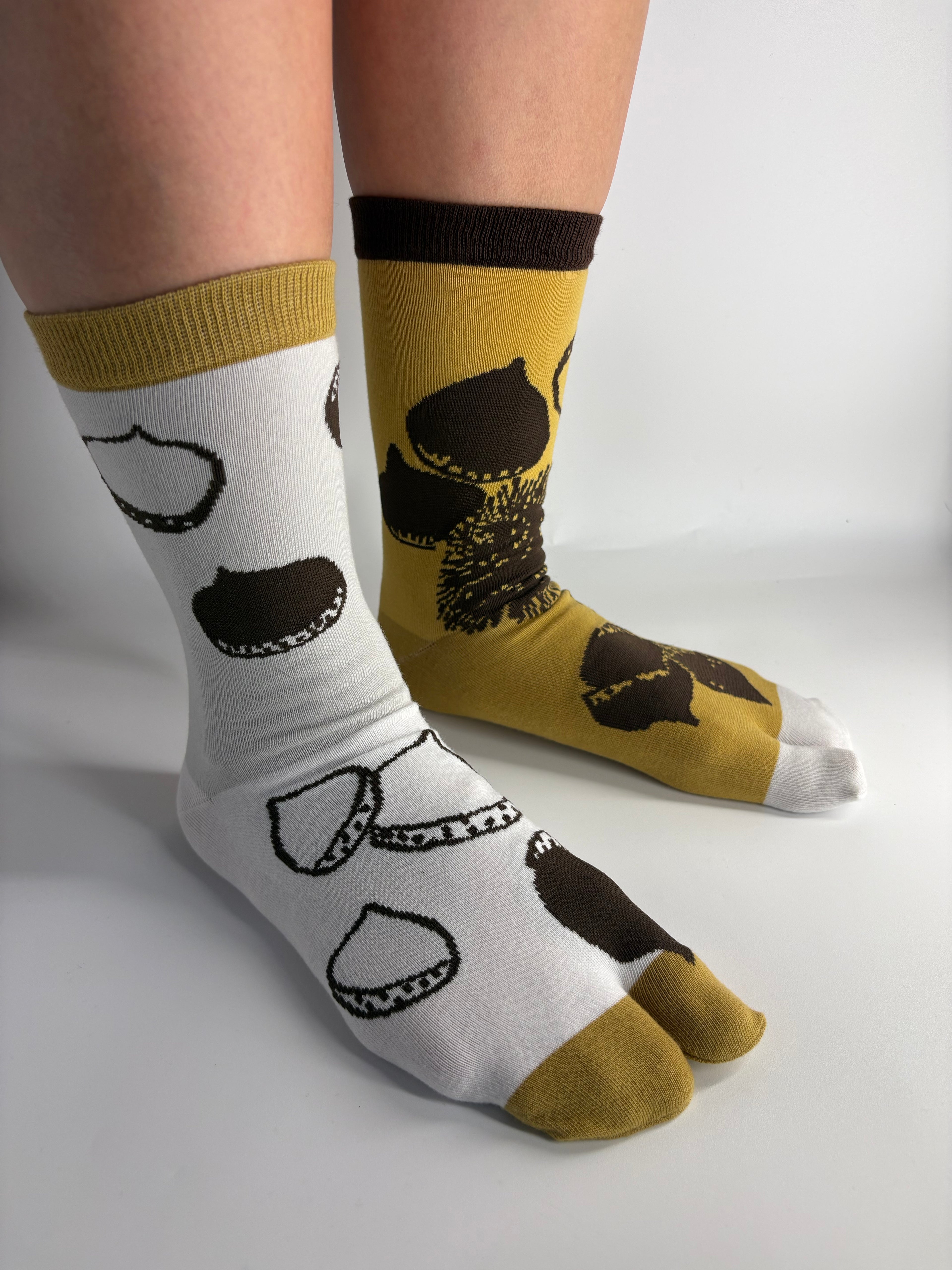 A pair of split-design socks worn on feet, with one sock being white and the other mustard yellow, both featuring dark brown achestnut illustration.