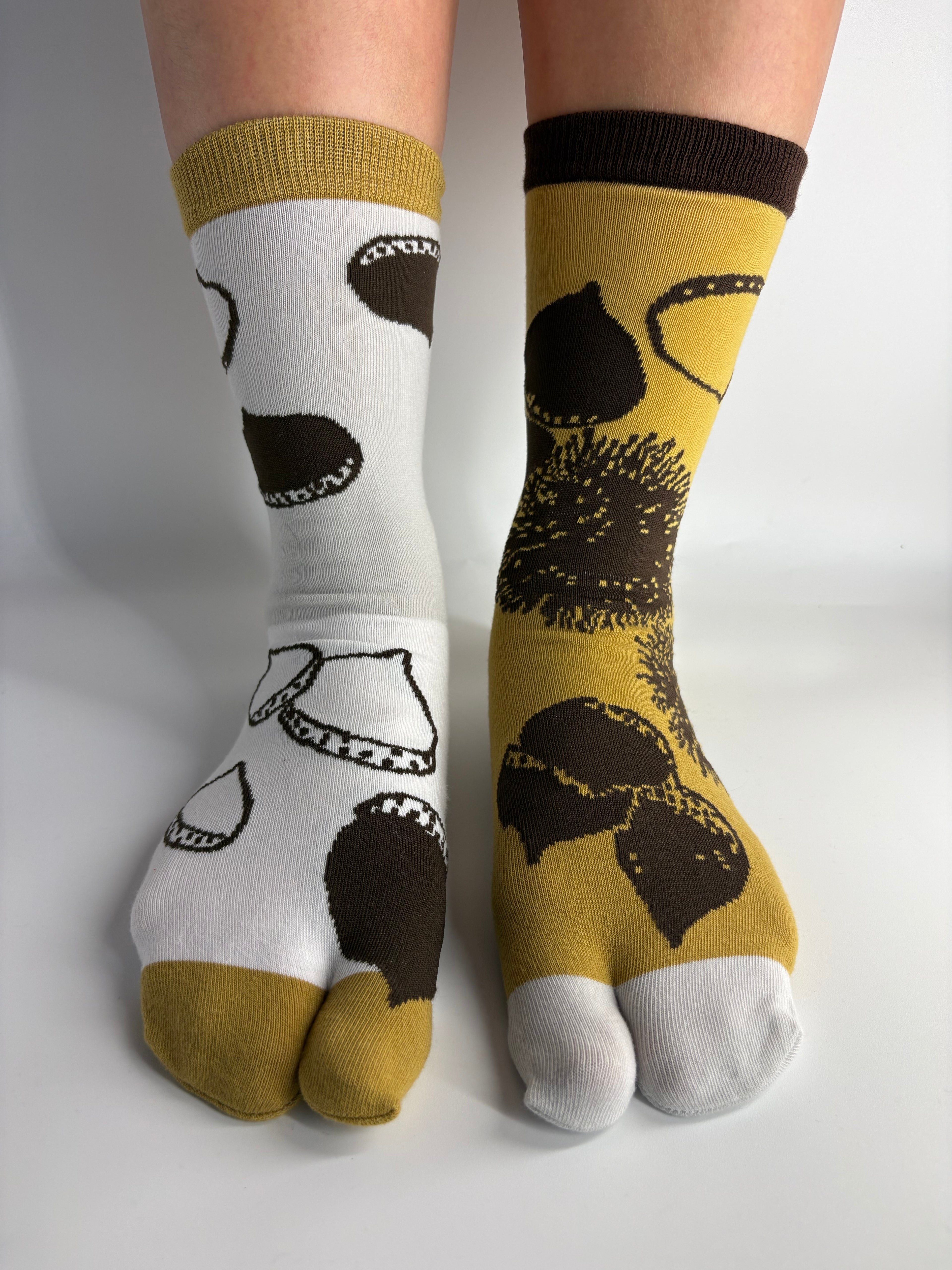 A pair of split-design socks worn on feet, with one sock being white and the other mustard yellow, both featuring dark brown achestnut illustration.