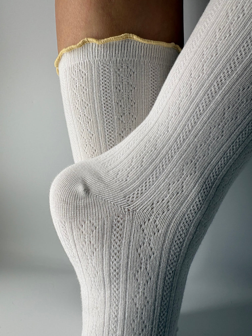 A close-up of a person's foot and lower leg wearing a pair of light white pointelle ribbed knit socks with a scalloped yellow trim at the top.