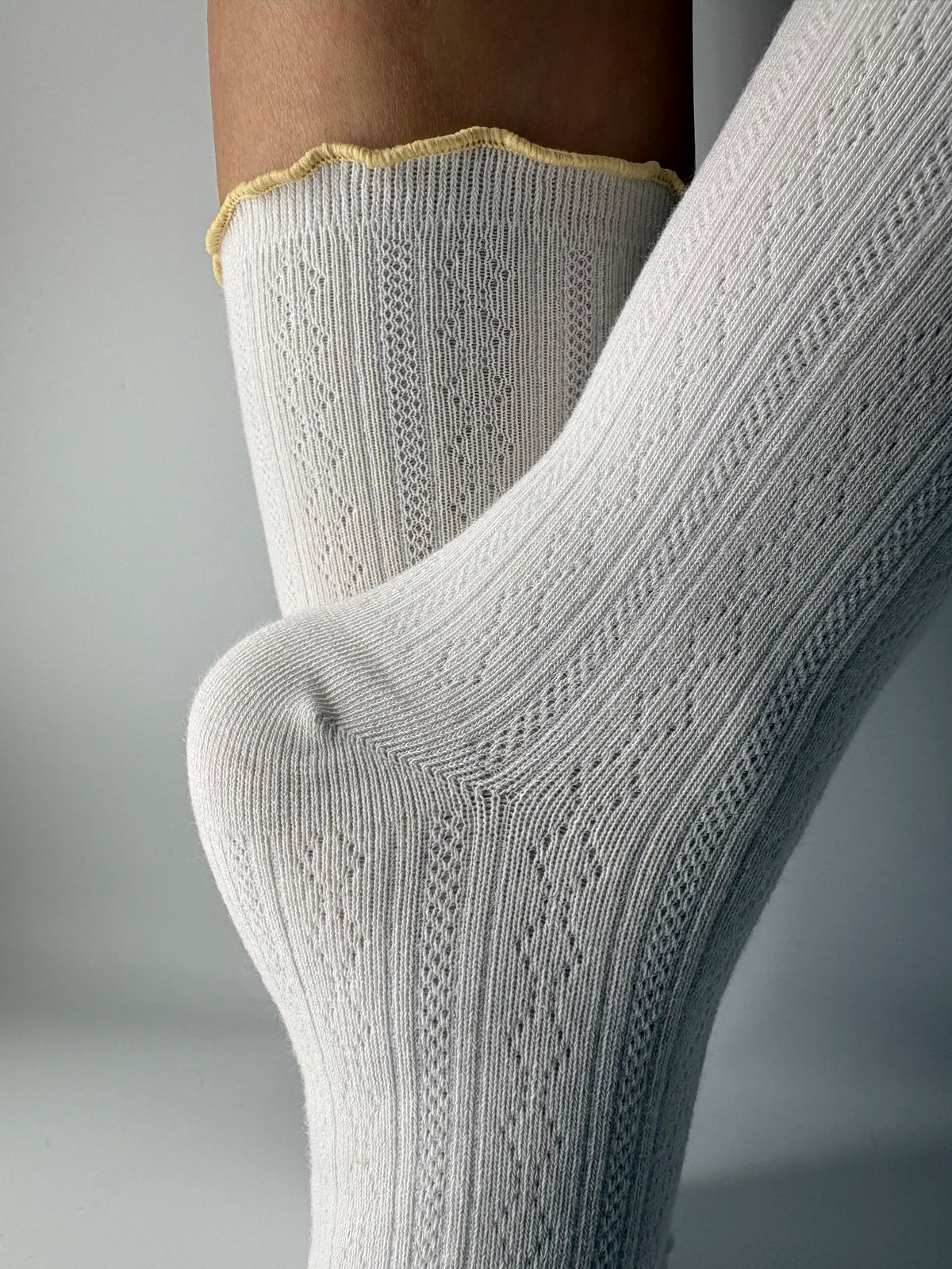 A close-up of a person's foot and lower leg wearing a pair of light white pointelle ribbed knit socks with a scalloped yellow trim at the top.