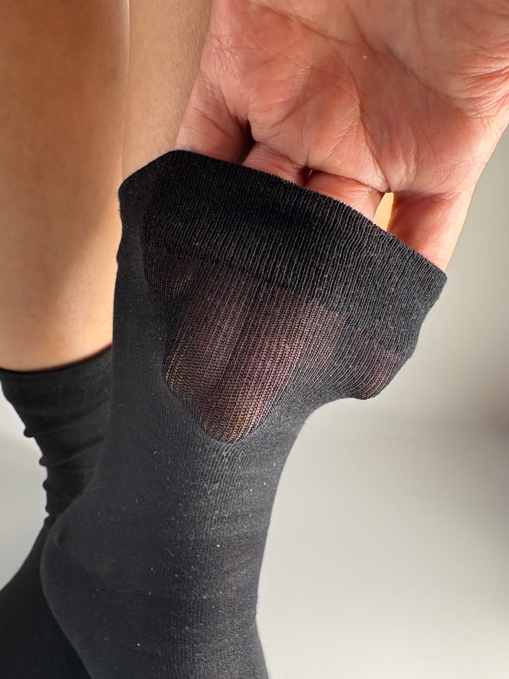 A close-up of a person's foot wearing a black sheer tabi sock, with a hand lifting the cuff to show the sheer mesh fabric on the cuff
