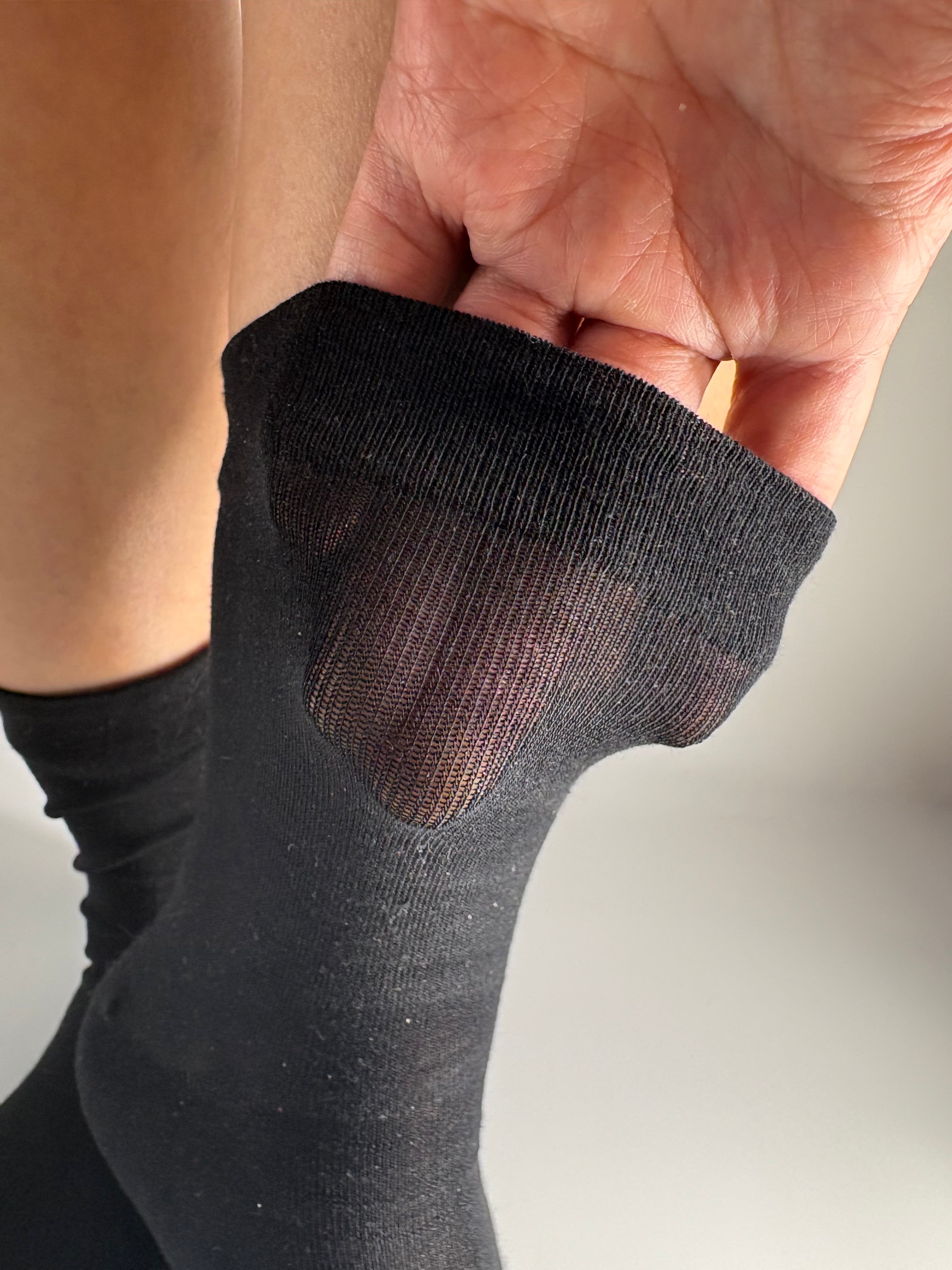 A close-up of a person's foot wearing a black sheer tabi sock, with a hand lifting the cuff to show the sheer mesh fabric on the cuff